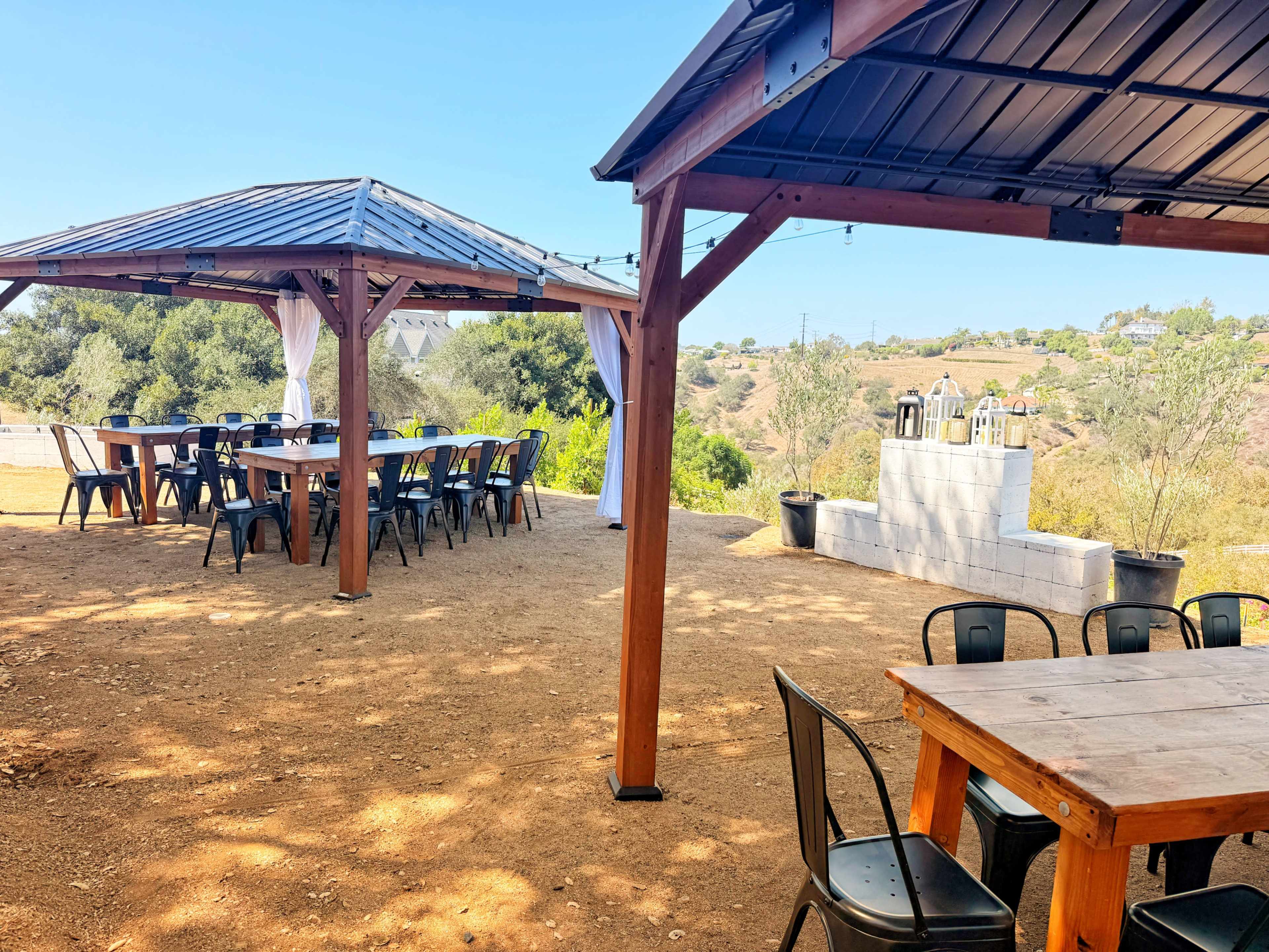 The image shows a rustic outdoor venue with two wooden pavilions and several tables set up on a dirt floor, surrounded by a scenic hillside.