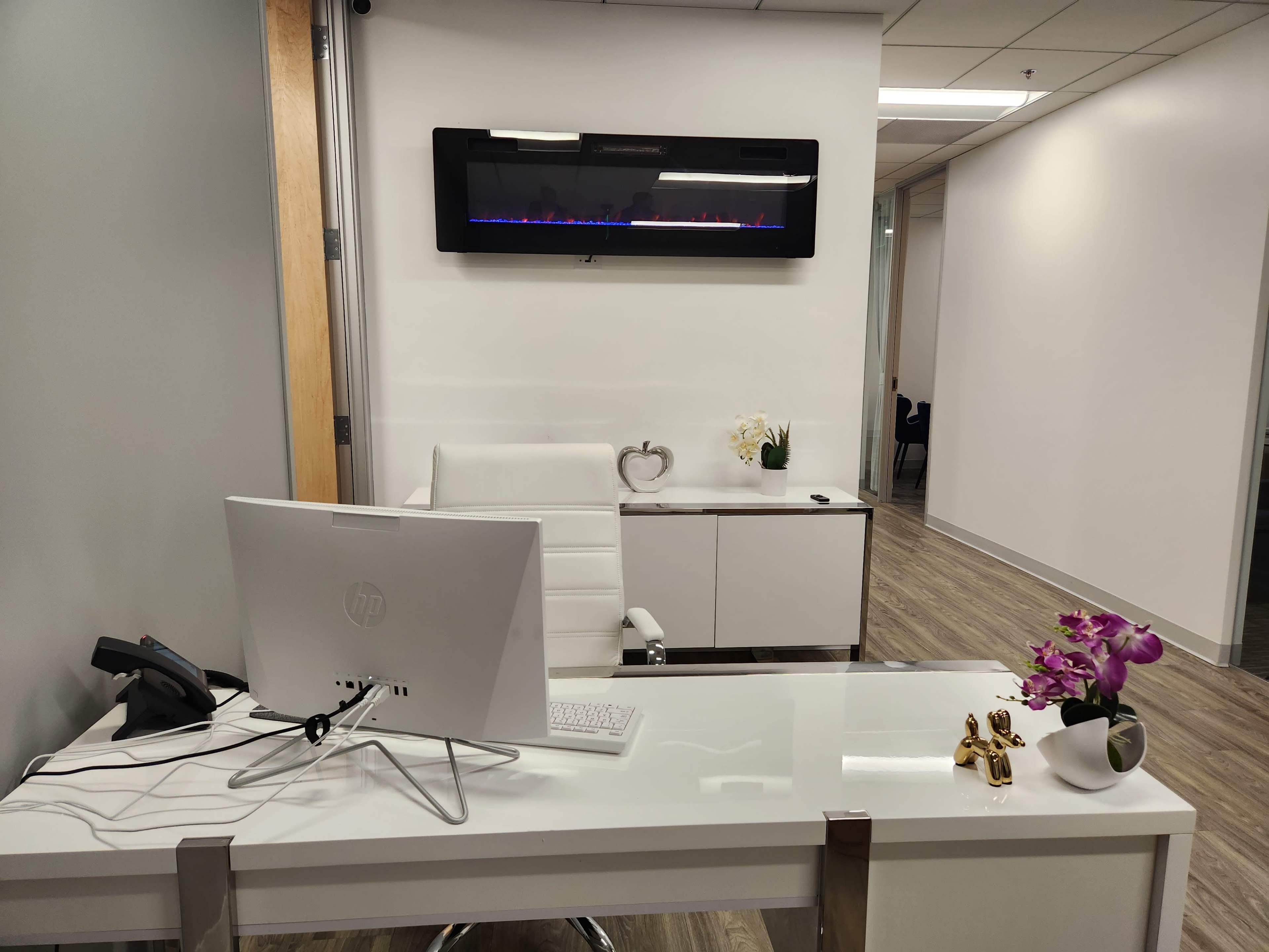 A modern office space featuring a white desk with a computer, a sleek chair, and a wall-mounted electric fireplace above a cabinet with decorative items.