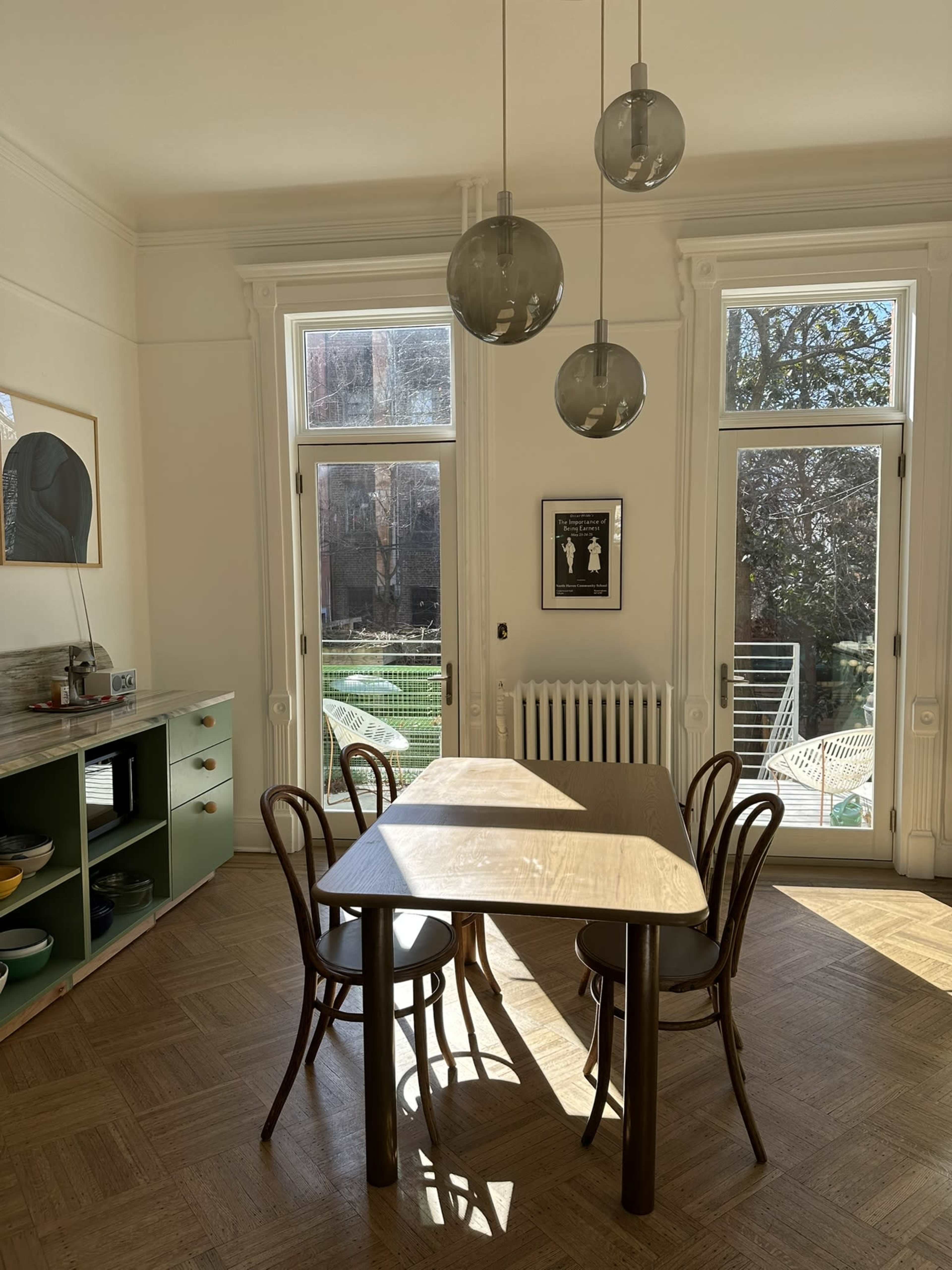 A dining area features a table surrounded by four chairs, with large windows letting in natural light and a view of an outdoor garden.