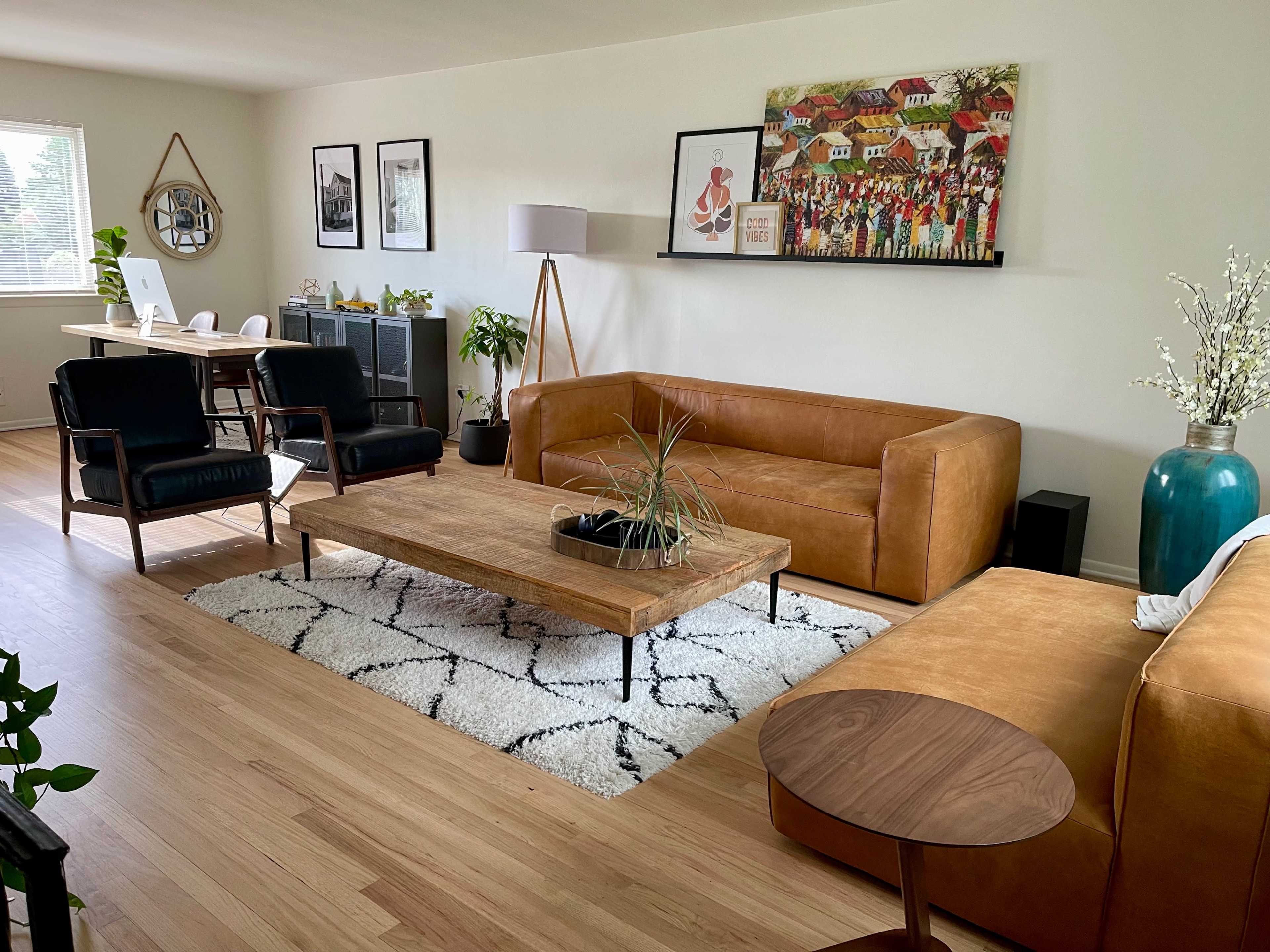 The living room features a brown leather sofa, a wooden coffee table, and a large wall art piece above, with a modern dining area visible in the background.