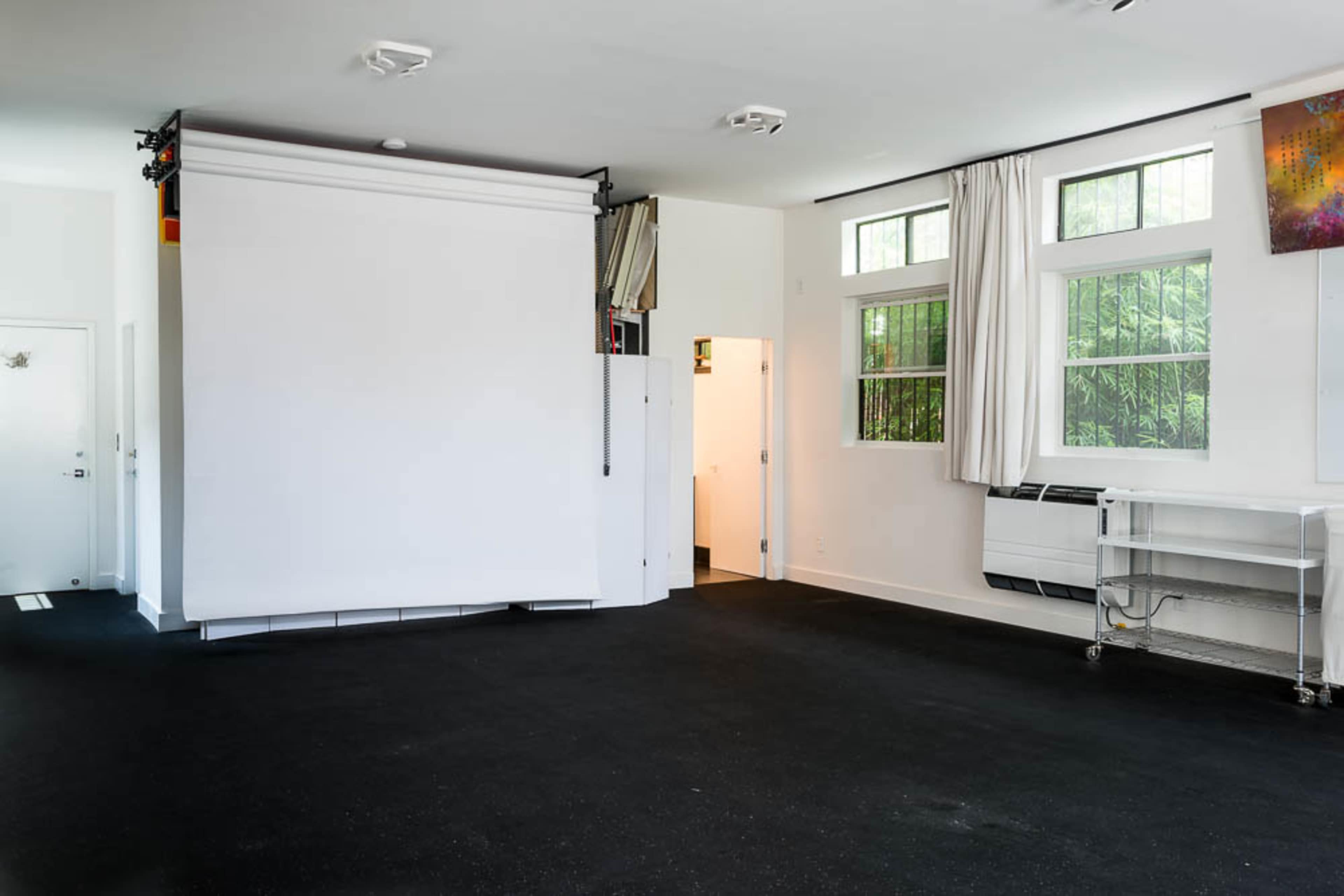 A minimalistic studio space with a retractable projector screen, large windows, and an entrance leading to another room.