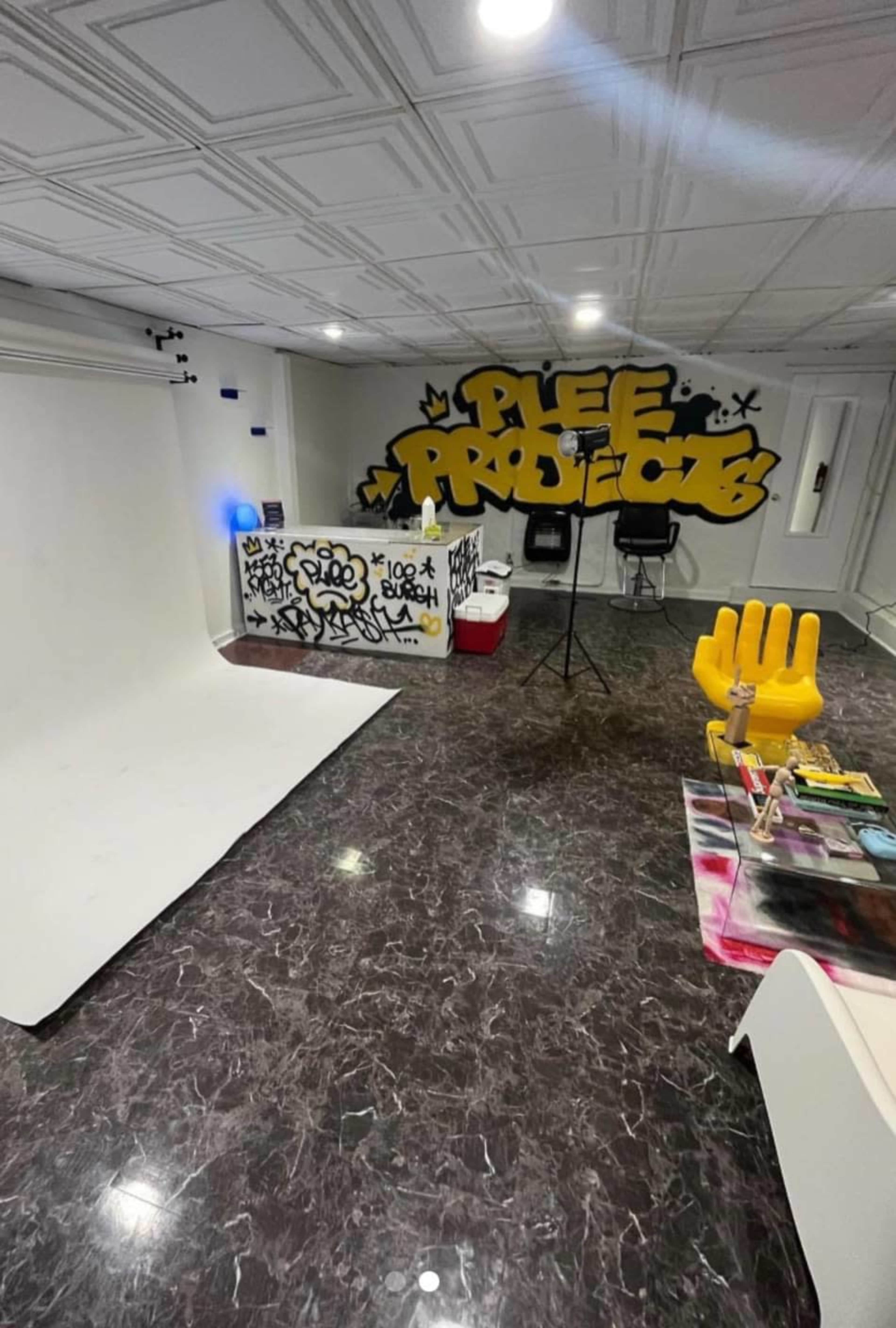A studio space with a graffiti wall featuring the words "Plee Projects," a stark white backdrop, and various artistic supplies arranged on a table.