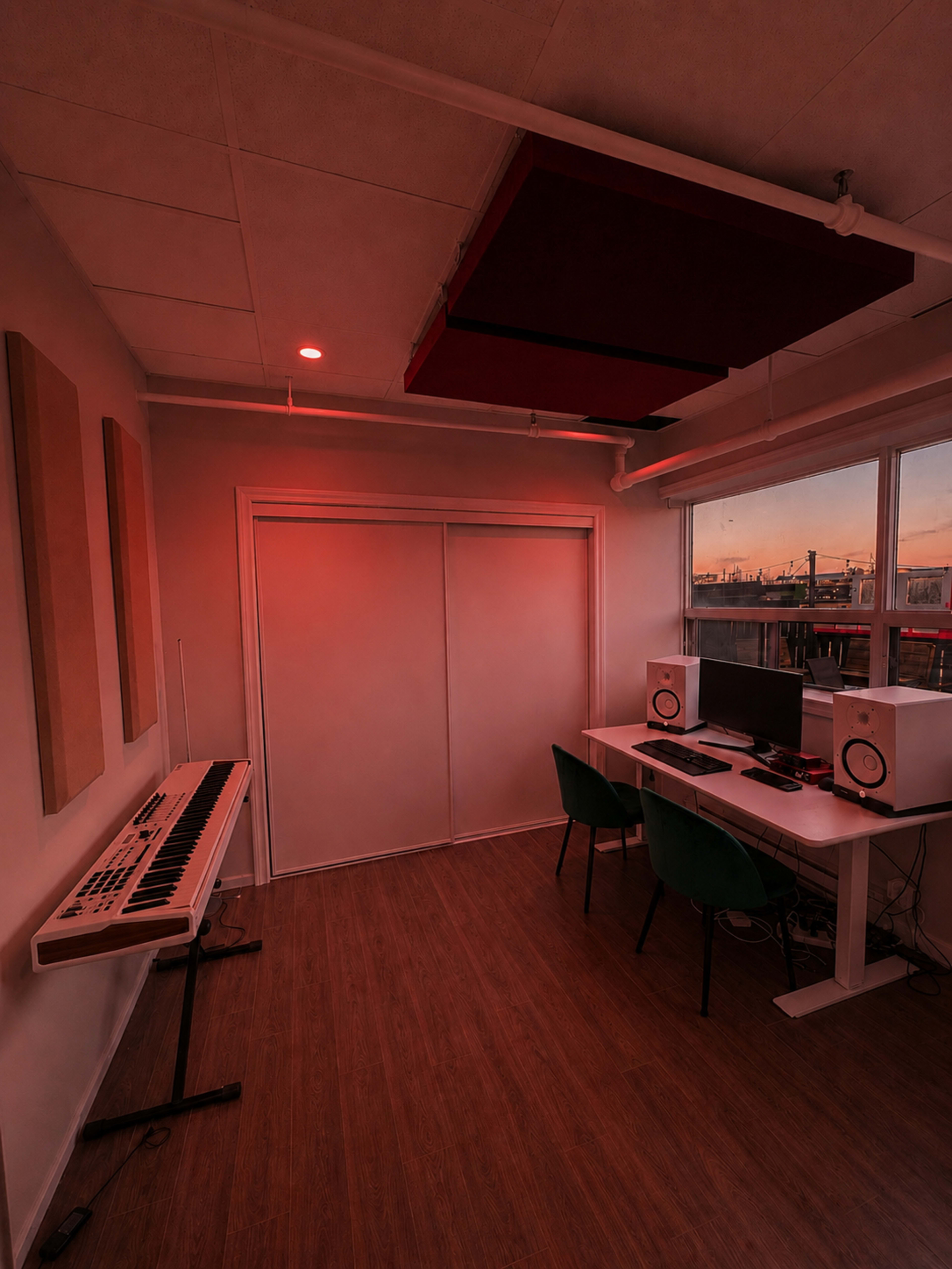 The image shows a music production studio with a keyboard, two monitors, and chairs, illuminated by red light near windows displaying a sunset.