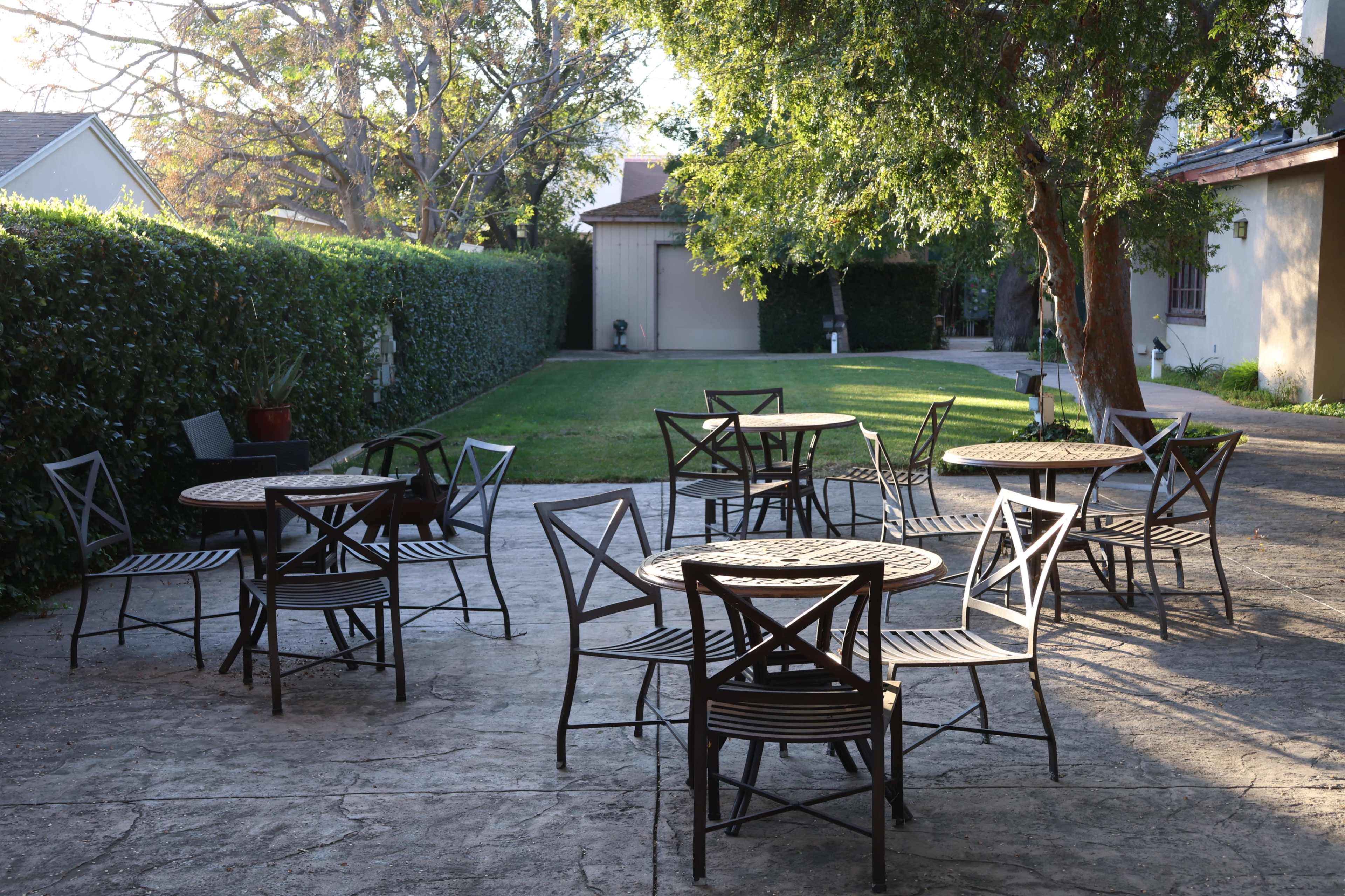 The image shows a concrete patio with several empty tables and chairs arranged under the shade of trees, surrounded by greenery.