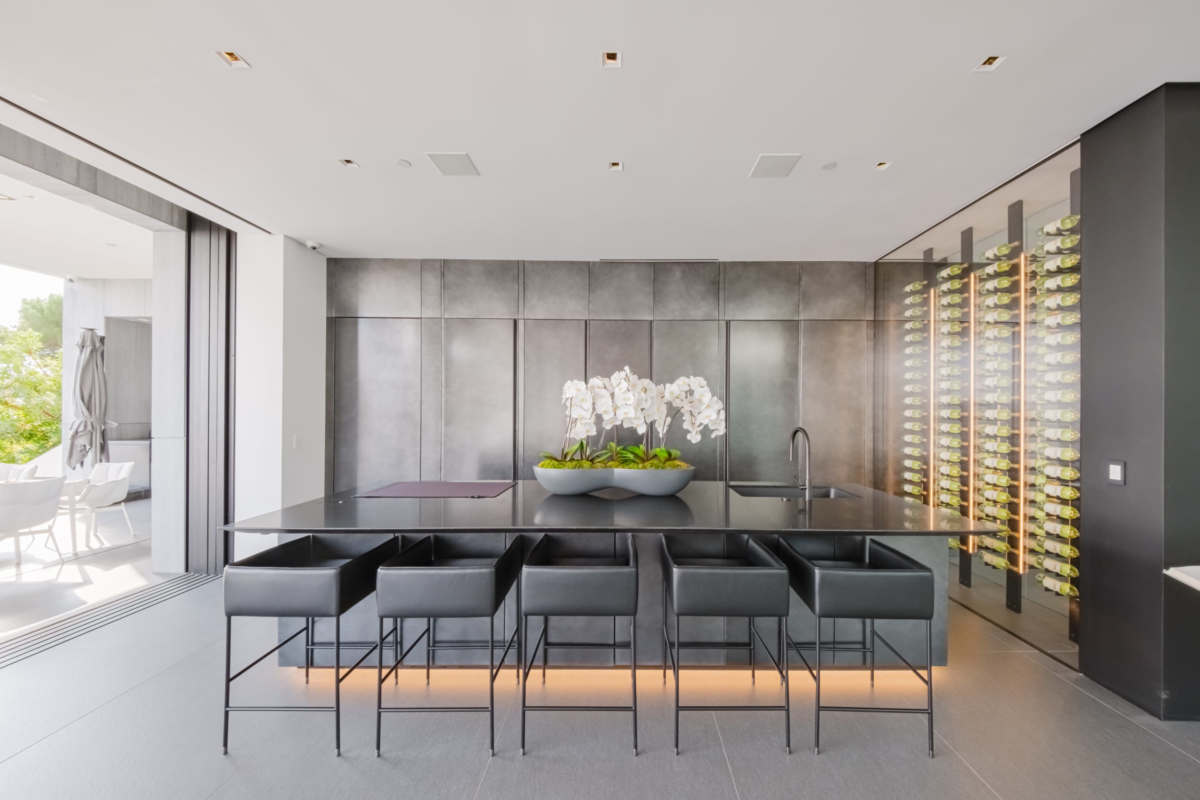 The image shows a modern kitchen with a sleek black island, five black bar stools, and a wall of wine storage beside a large window with outdoor seating.