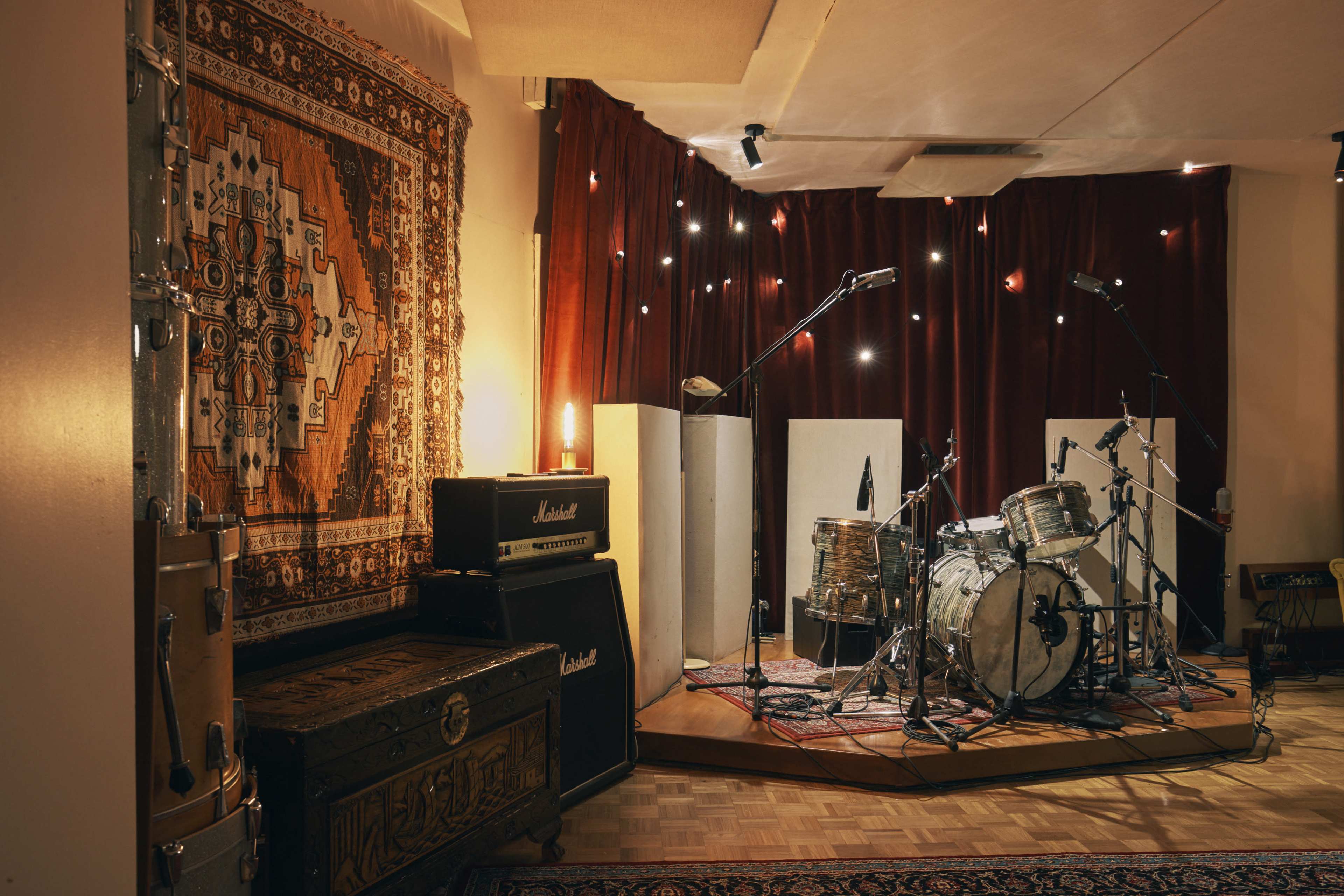 A recording studio features a drum set surrounded by soundproof panels and vintage amplifiers, with decorative rugs on the floor and walls.