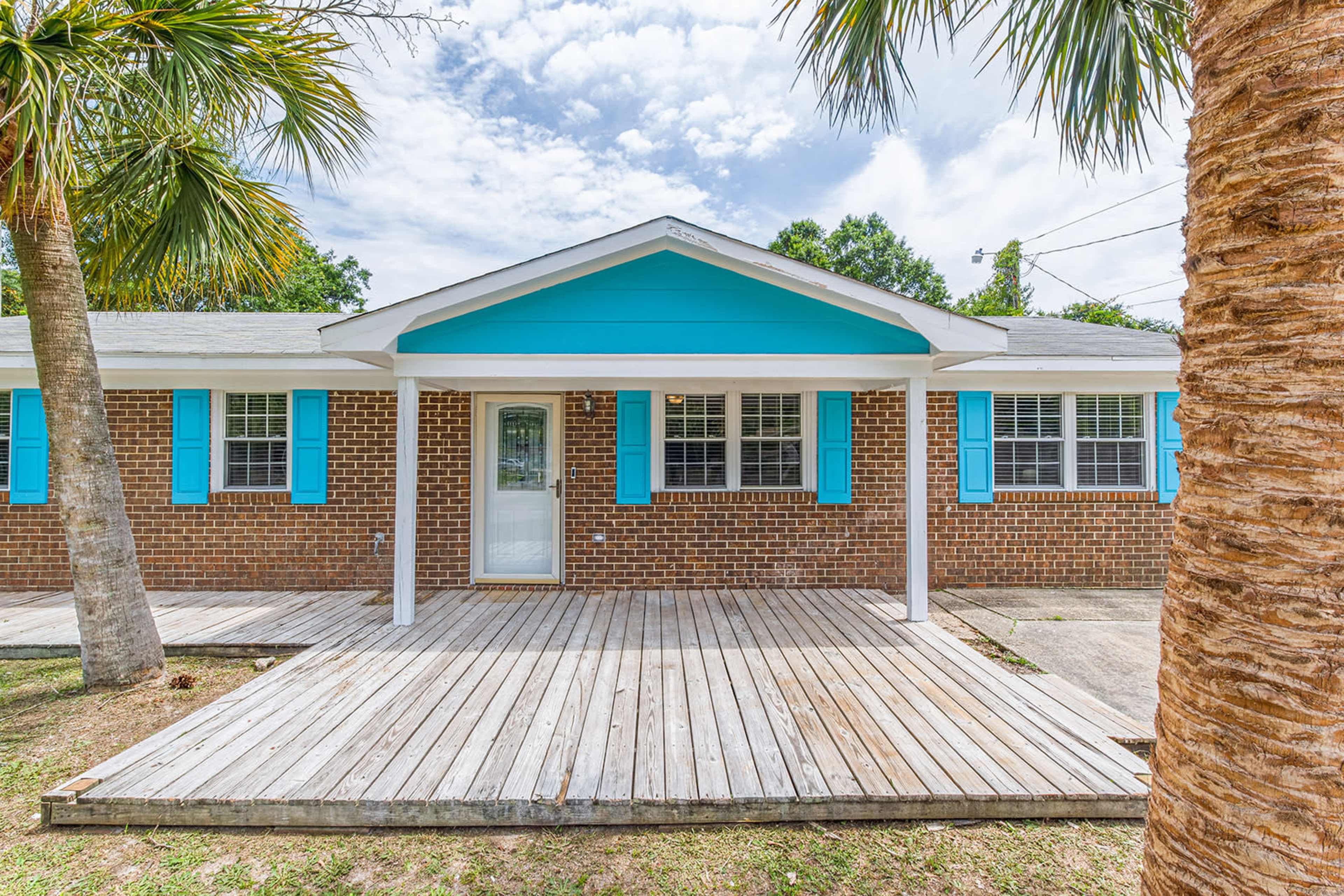 A brick house with turquoise shutters, a covered porch, and a wooden deck surrounded by palm trees.
