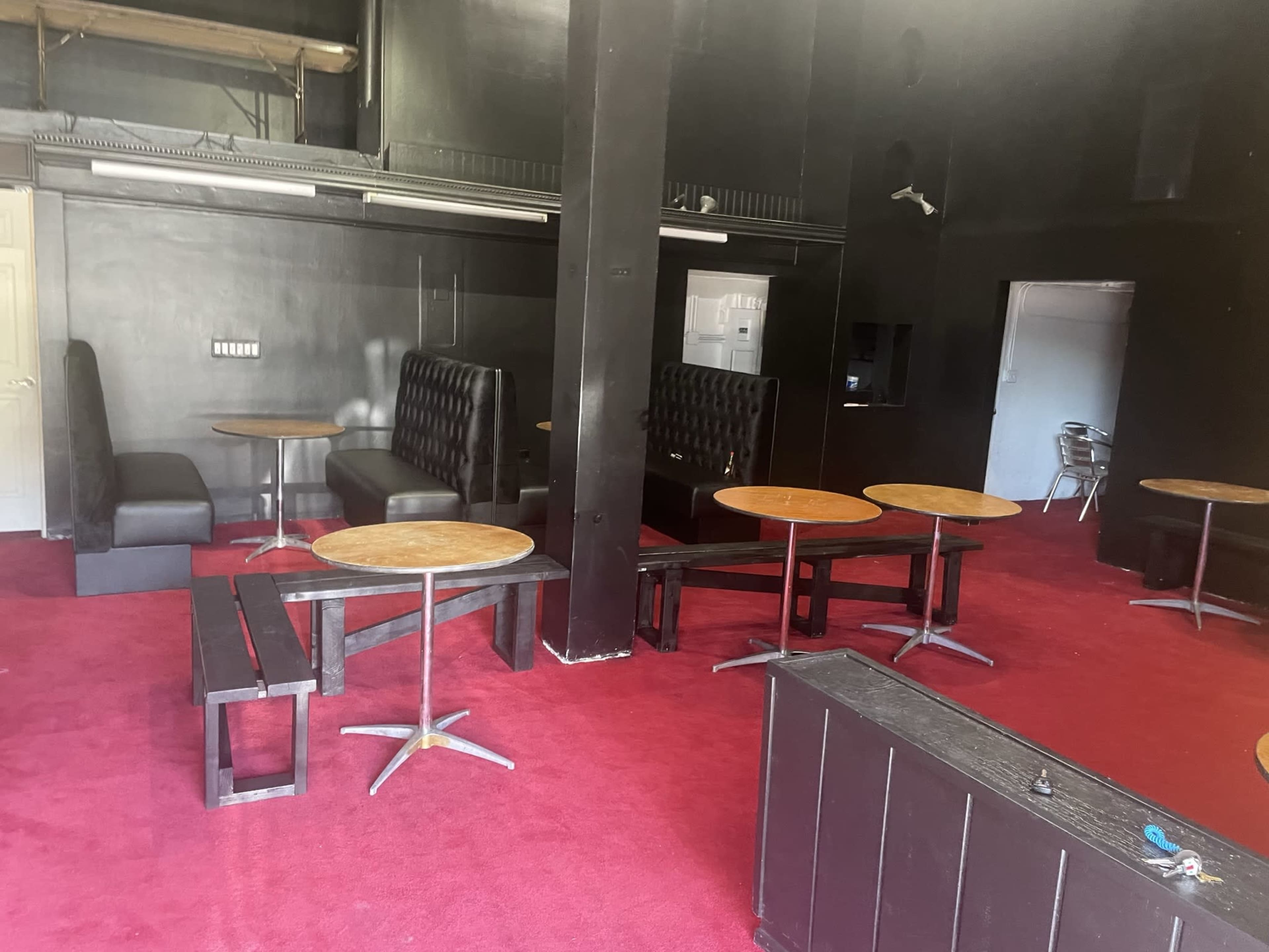 A dark room with red carpet, featuring several round tables and black booth seating arrangements.