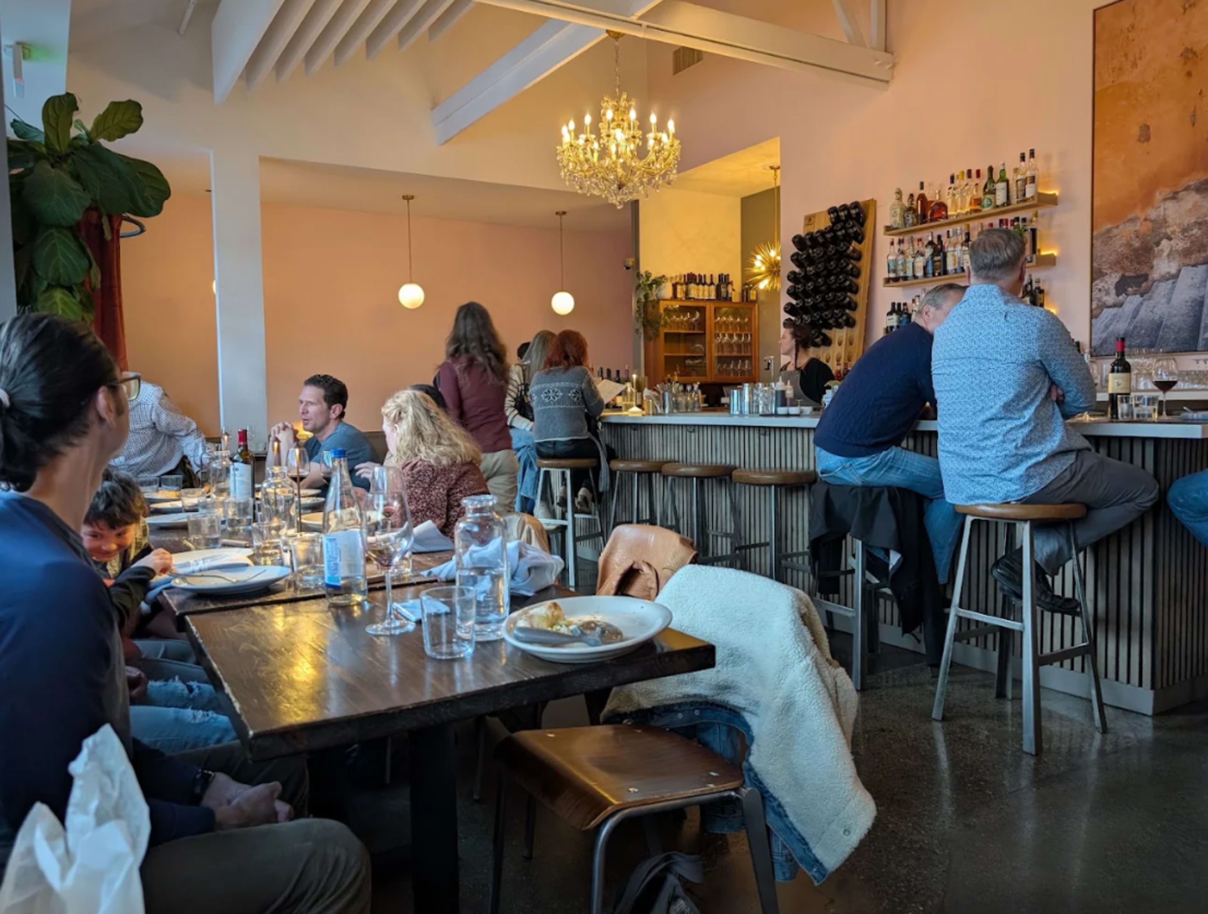 The image depicts a busy restaurant interior with patrons seated at tables and a bar, featuring a chandelier and natural light from large windows.
