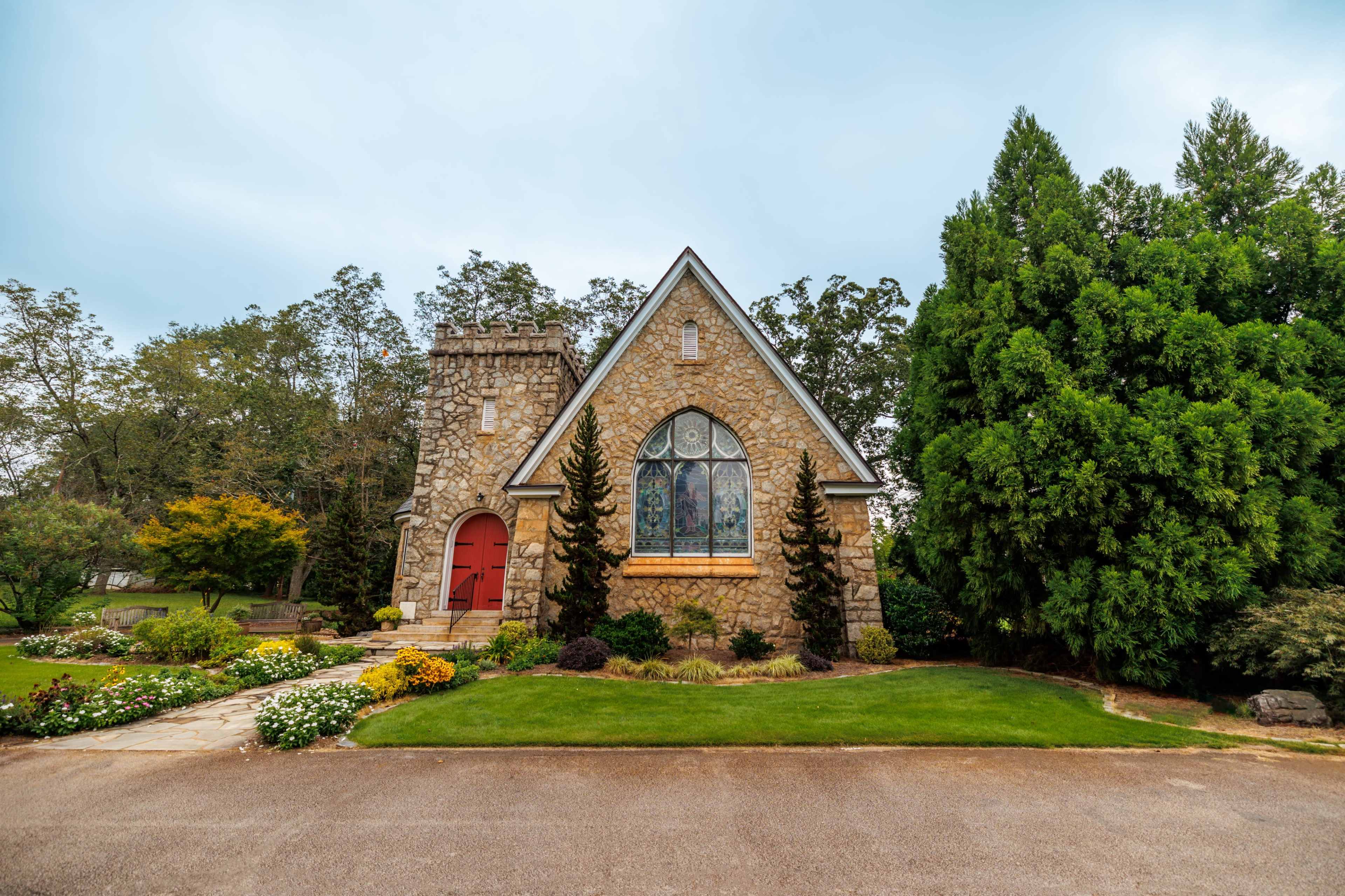 A stone church with a tall tower and stained glass windows is surrounded by well-maintained gardens and trees.