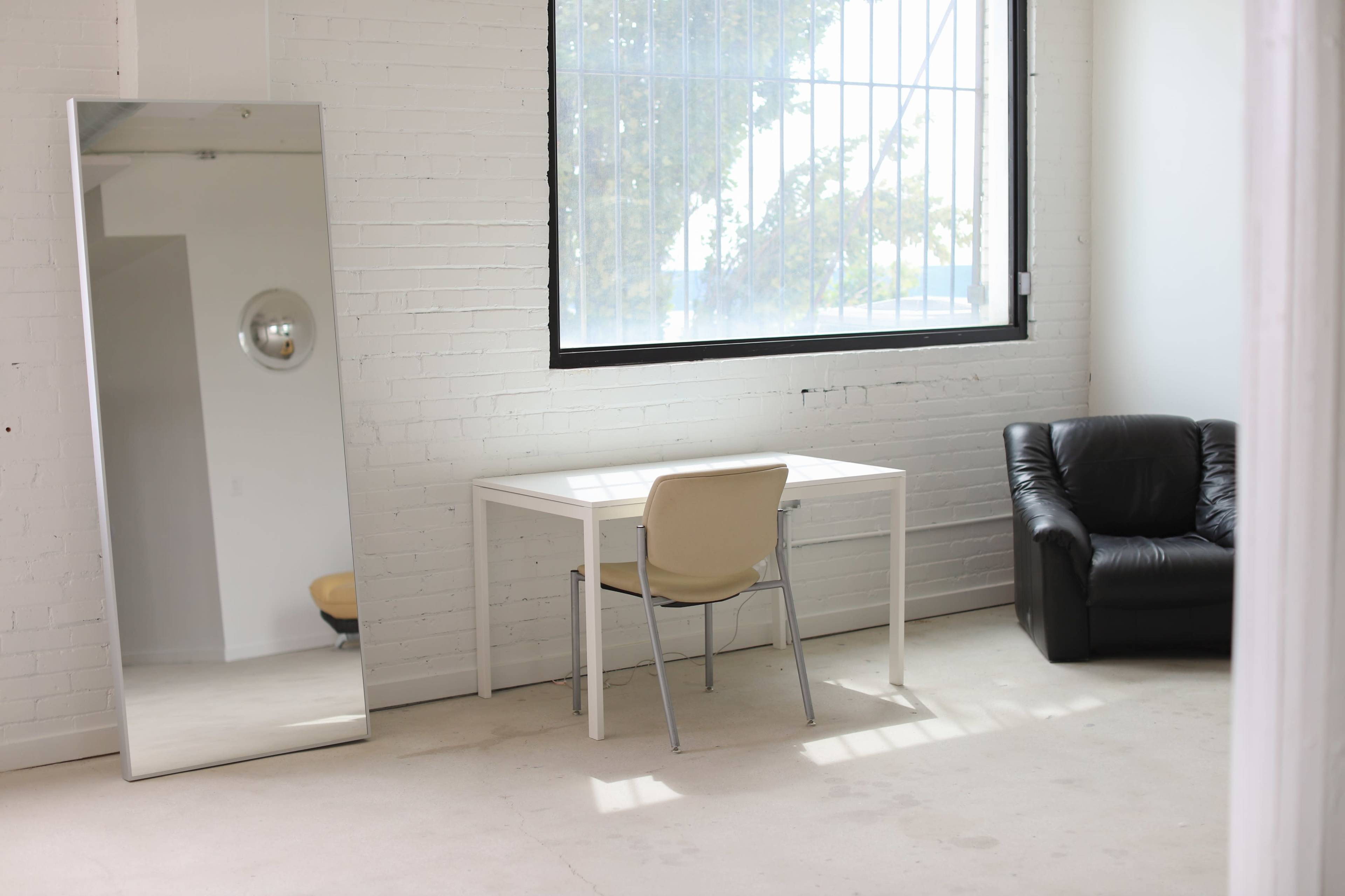 A modern room features a large mirror, a white desk with a chair, and a black leather armchair beside a window.