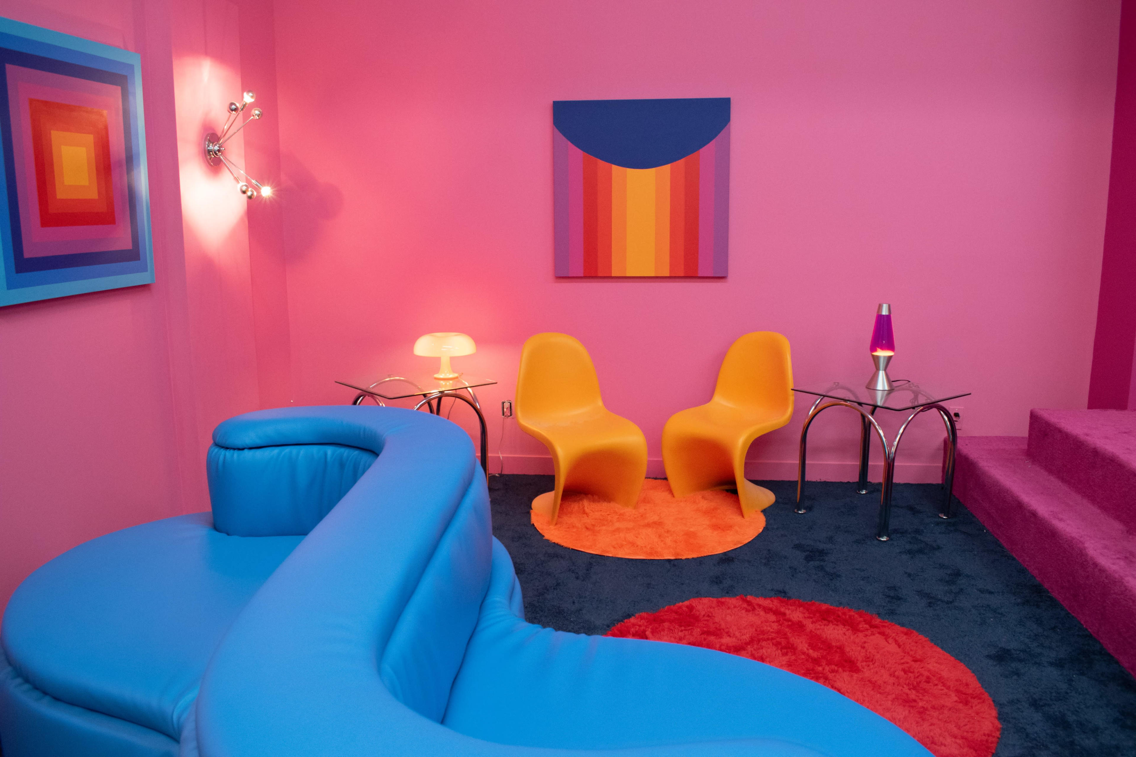 The room features a pink wall, a blue curved sofa, two orange chairs, a colorful wall art piece, and a small lamp on a glass table.