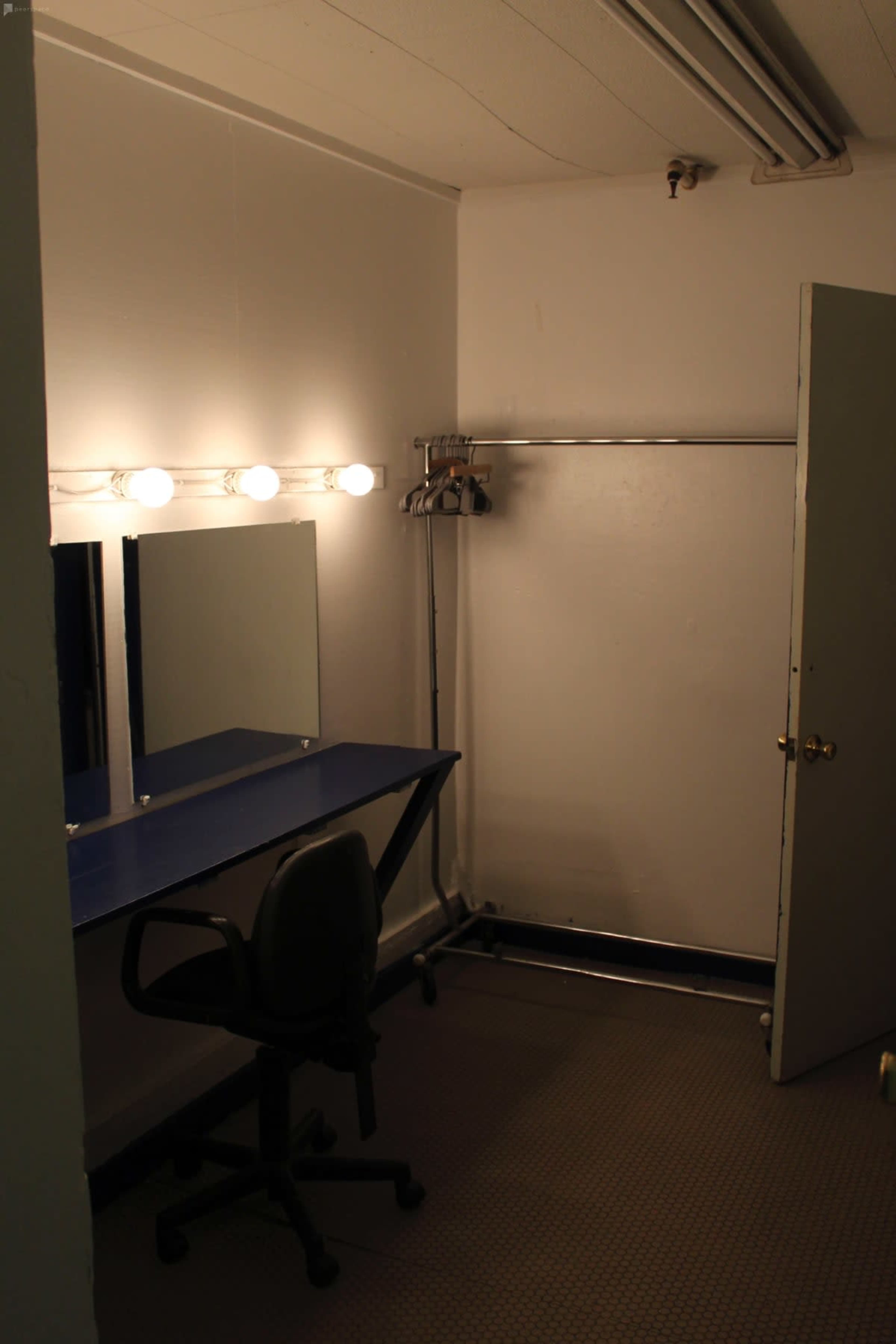 The image shows a small, dimly lit dressing room with a mirror, a blue countertop, a chair, and a clothing rack against the wall.