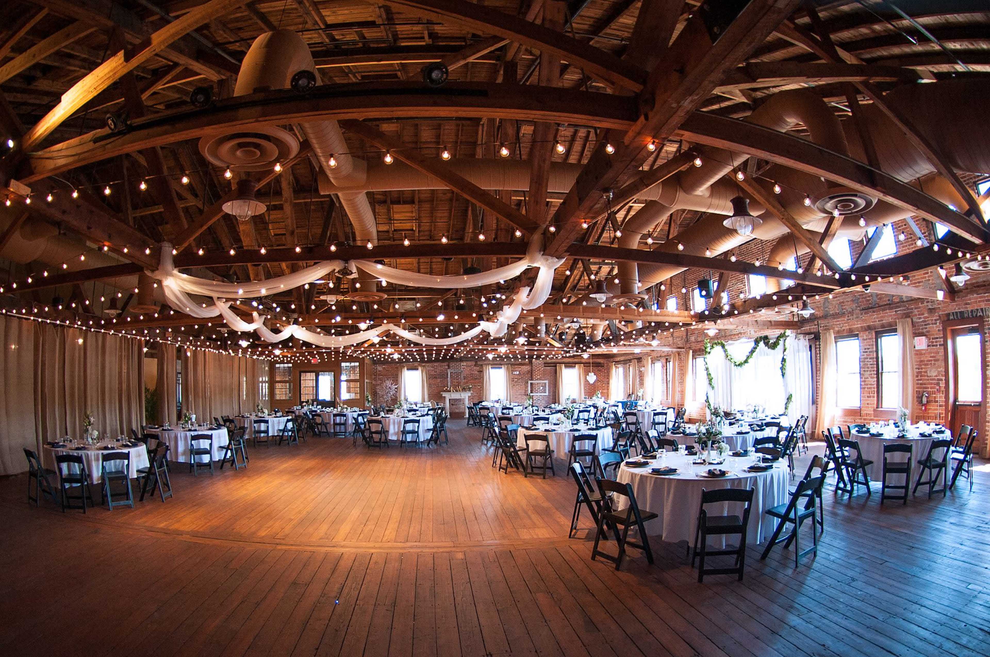 A large event space with multiple round tables covered in white tablecloths, set up under string lights and draped fabric.