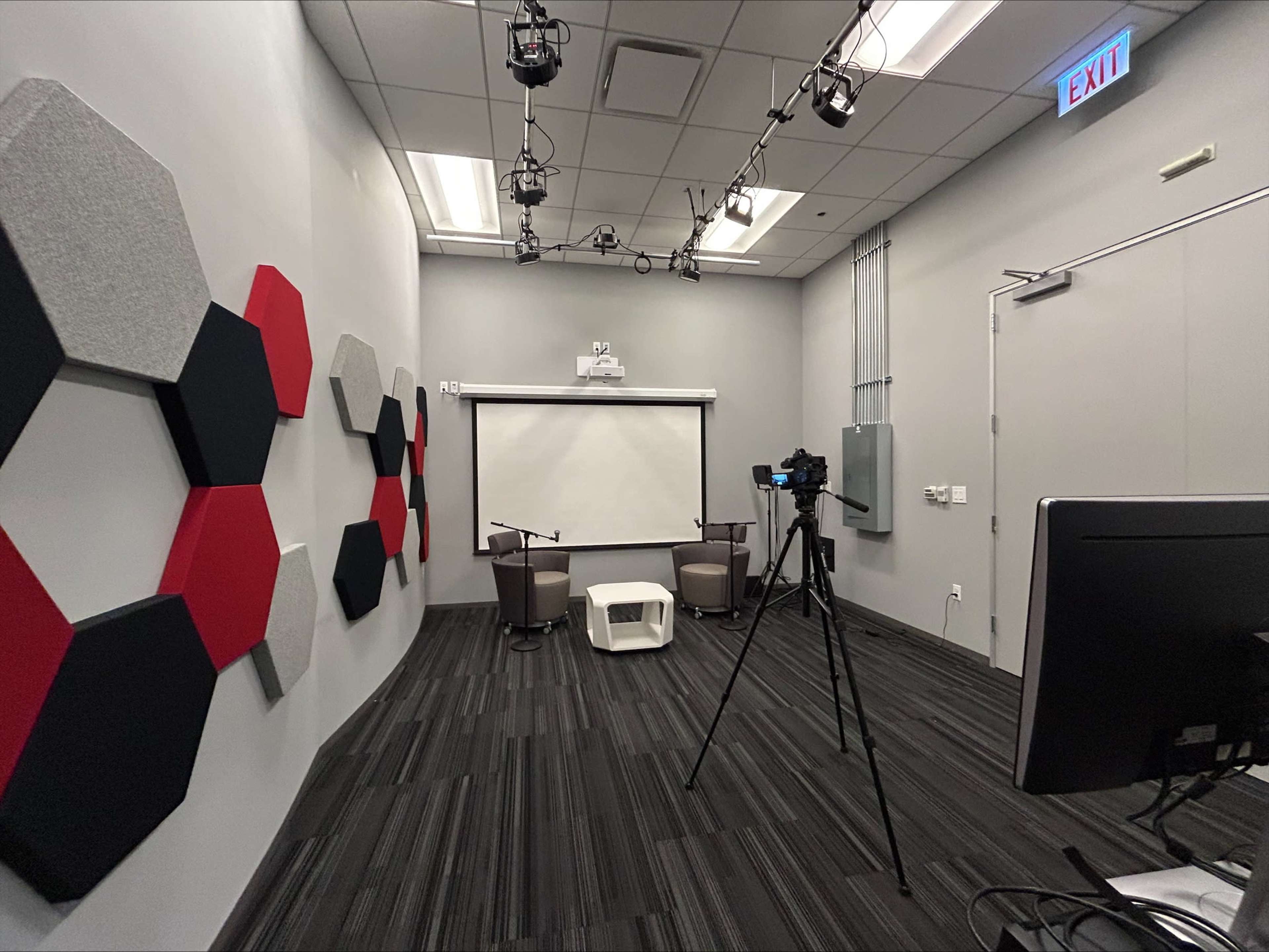 The image shows a modern studio setup featuring a camera on a tripod, a projector screen, and seating arrangements with hexagonal wall decorations.