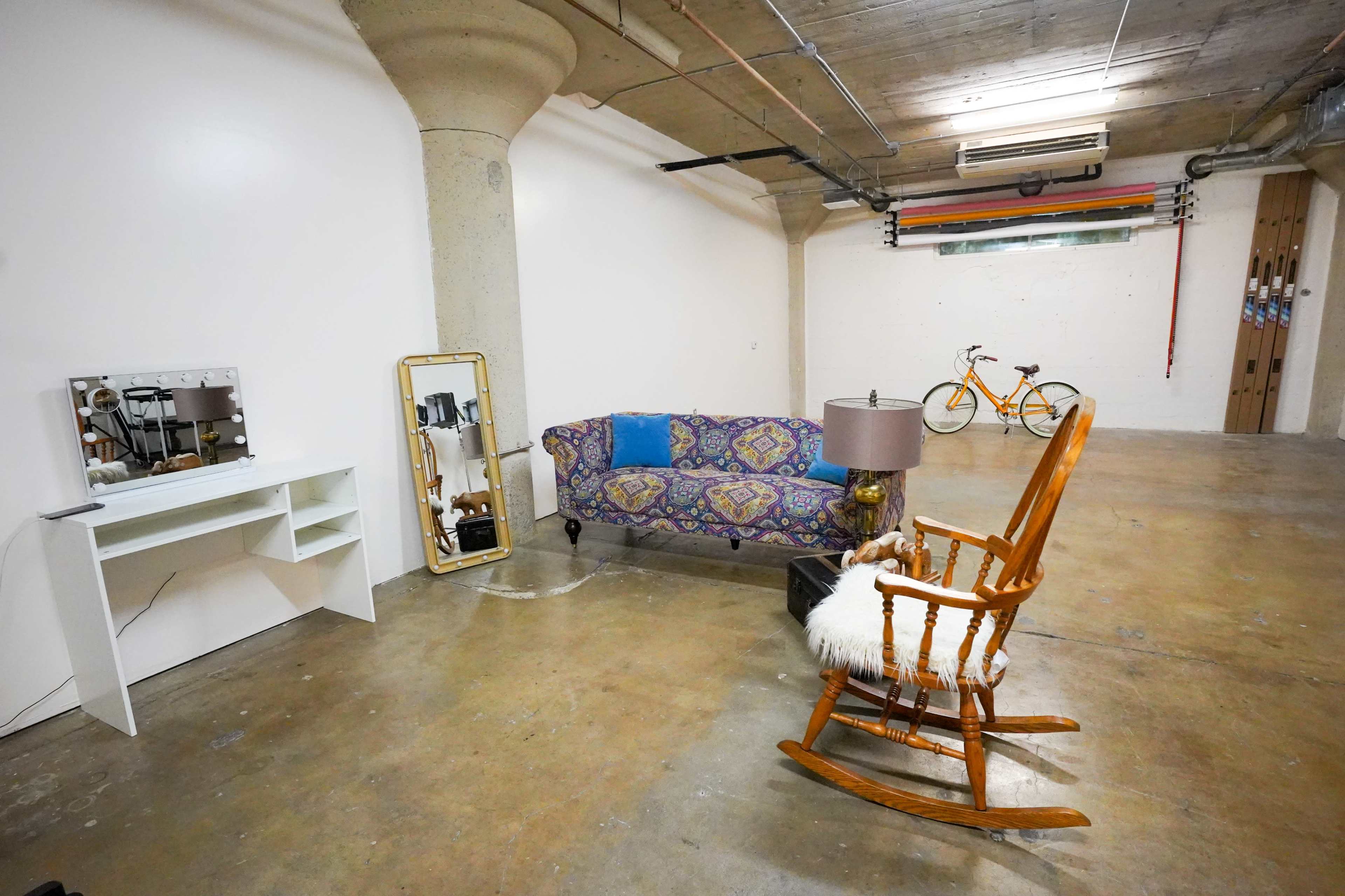 A spacious, unfinished room featuring a patterned couch, a rocking chair, a desk with a mirror, and a bicycle hanging on the wall.
