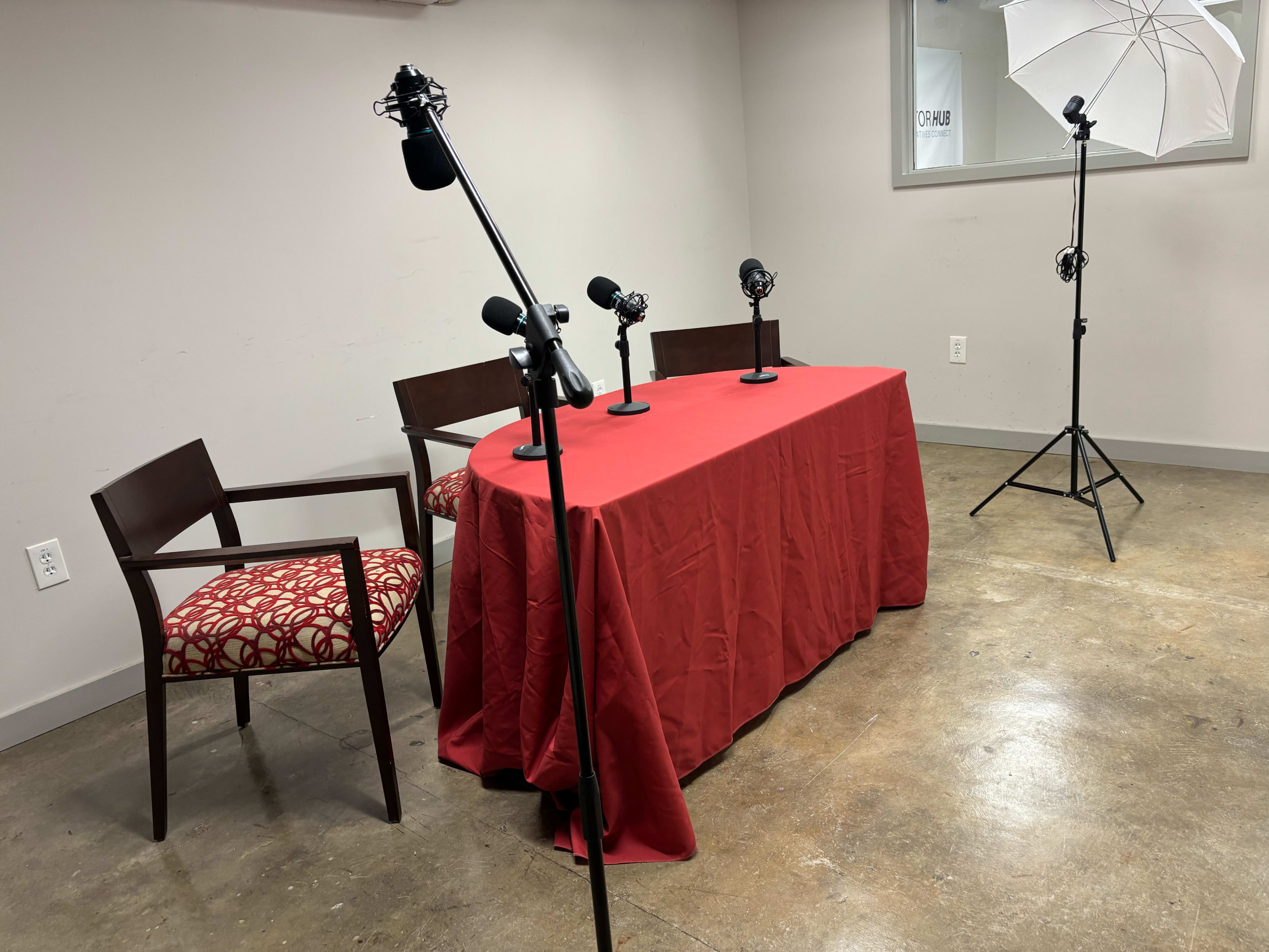 The image shows a small studio setup with a red table draped with a tablecloth, three microphones on stands, two wooden chairs, and a light umbrella positioned nearby.
