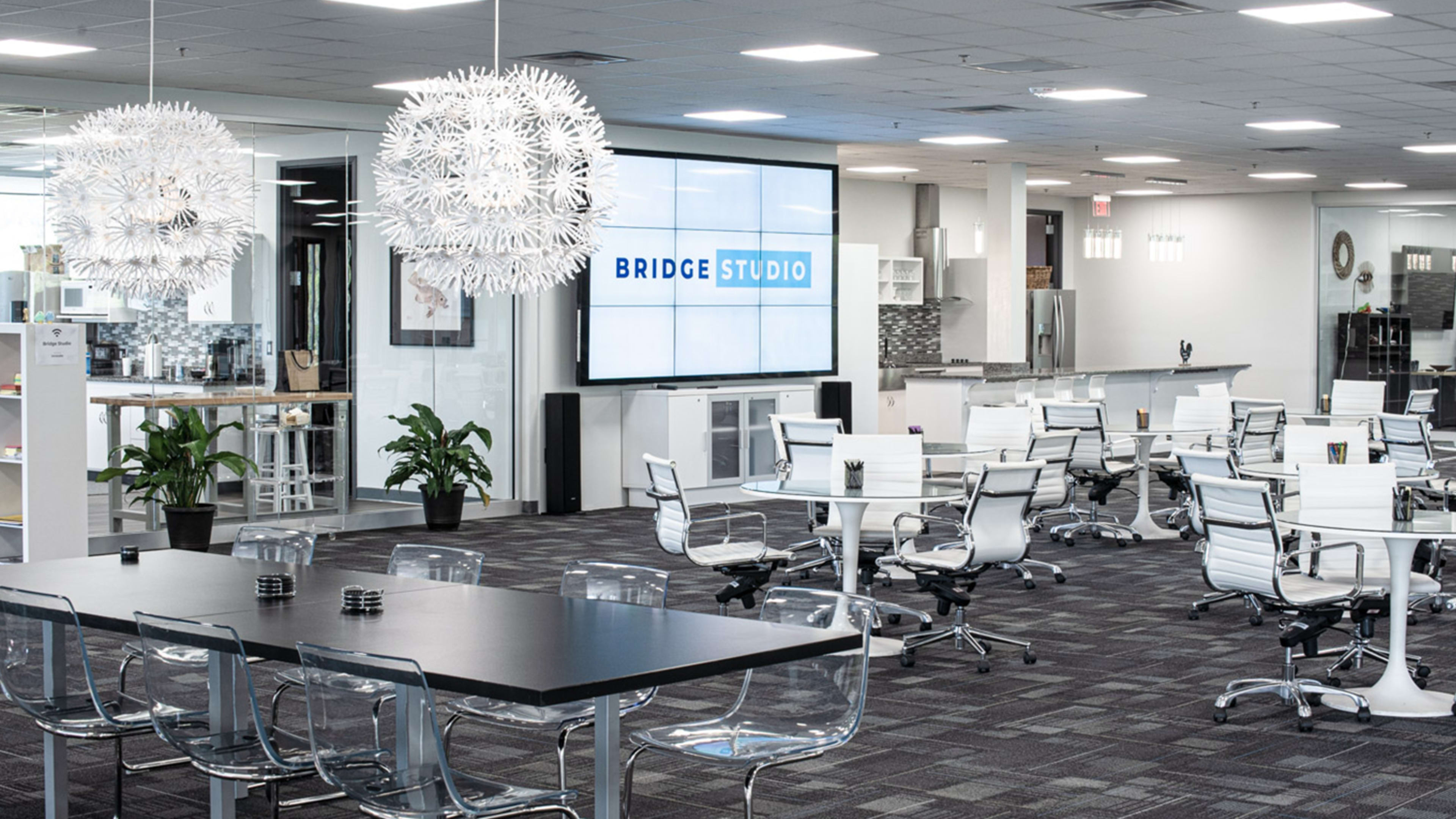 A modern office space with a large screen displaying "BRIDGE STUDIO," surrounded by transparent chairs and a kitchen area in the background.
