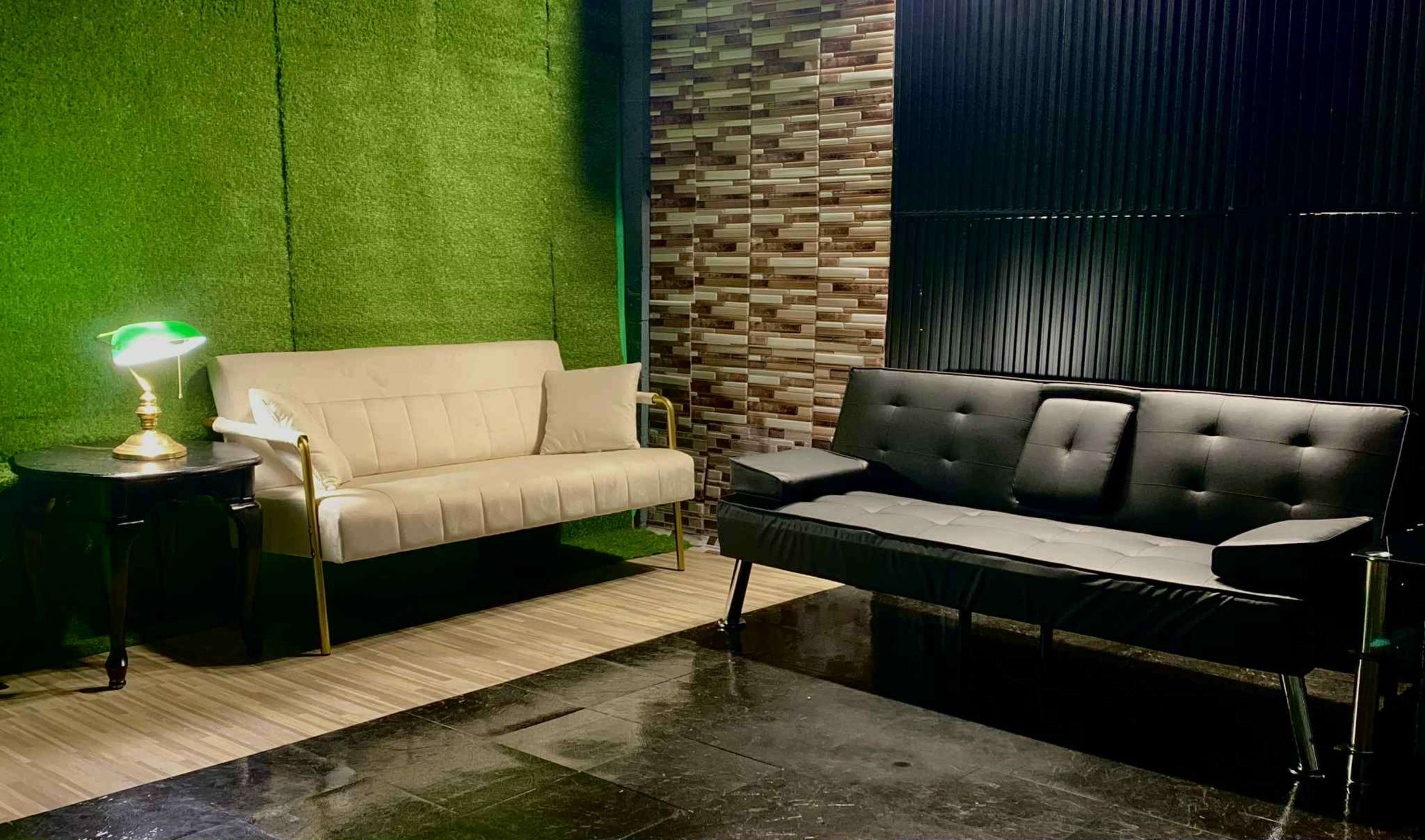 The image shows a modern seating area with a white sofa and a black sofa, accompanied by a decorative lamp on a small table against a backdrop of green grass and textured wall panels.