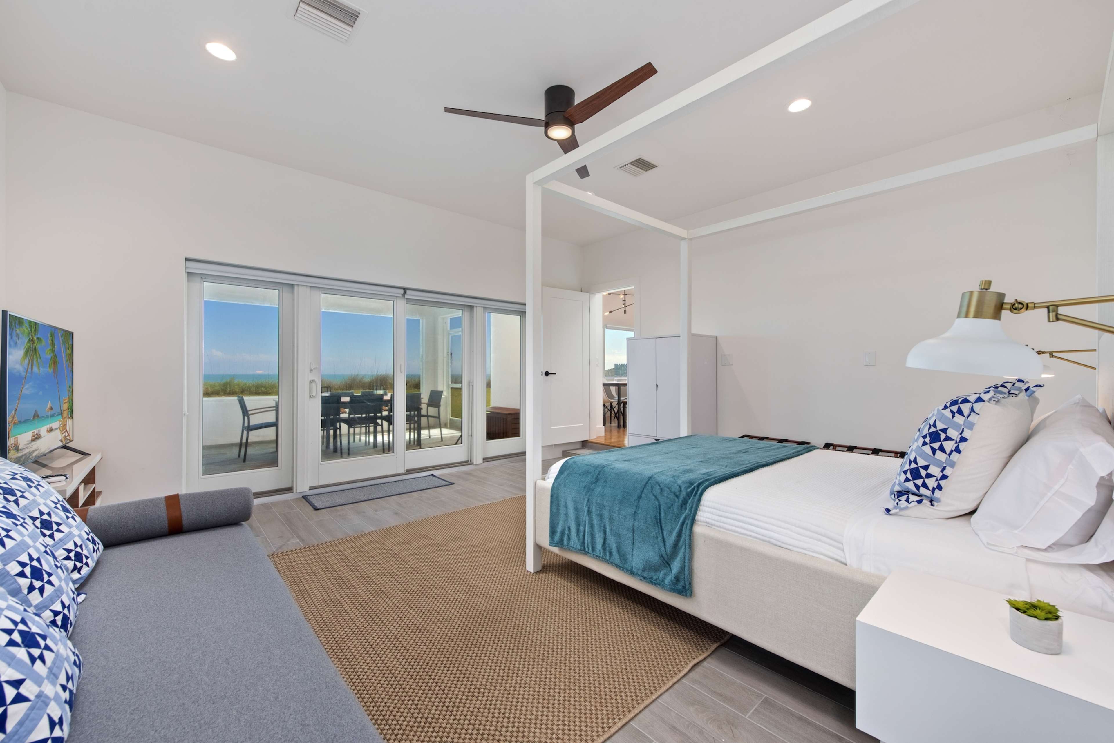 The image features a bright, modern bedroom with a four-post bed, a lounge area, and large sliding glass doors leading to an outdoor patio.