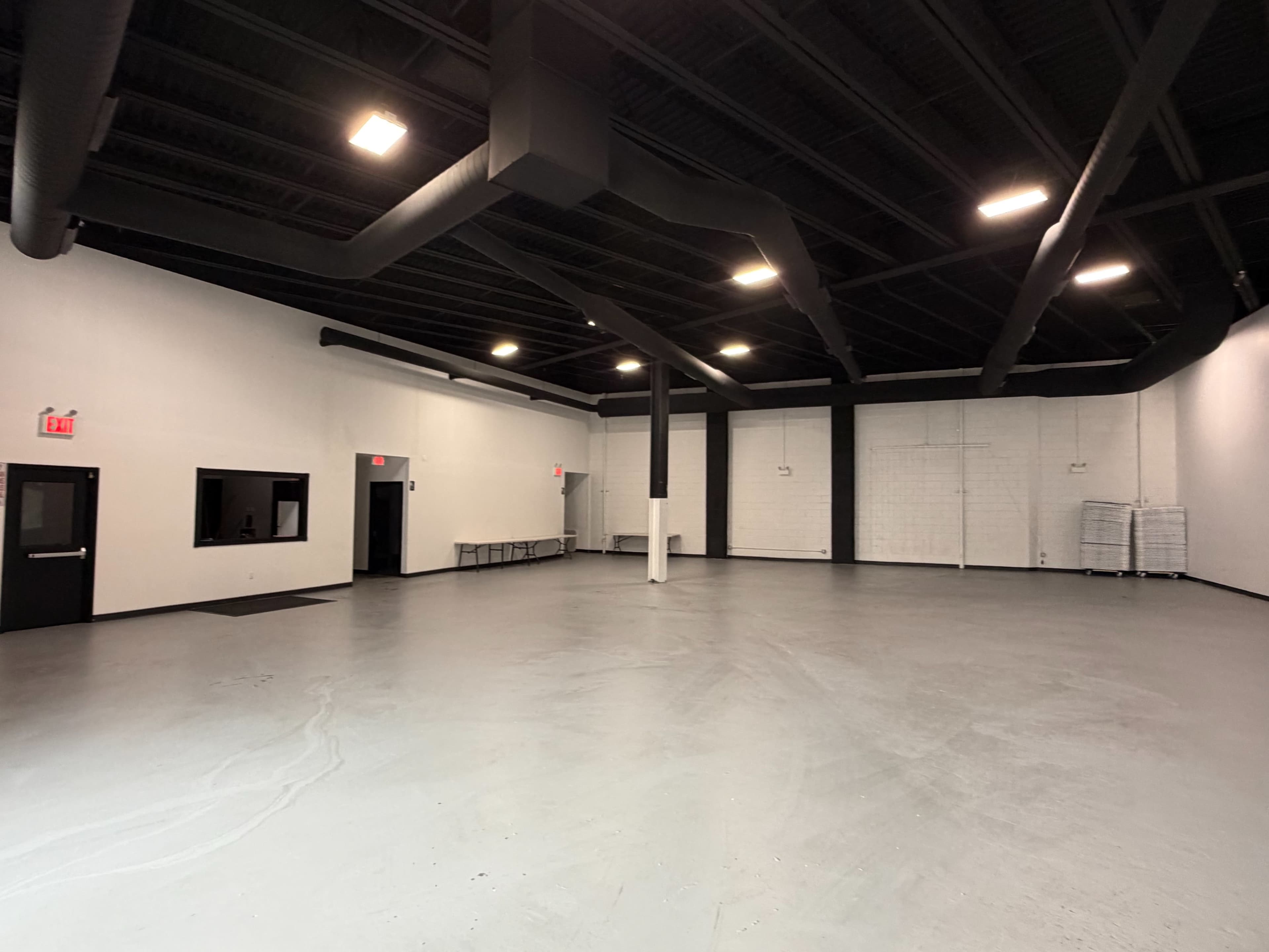 The image shows a large, empty commercial space with gray flooring and black-painted ceilings, featuring minimal furnishings and a few light fixtures.