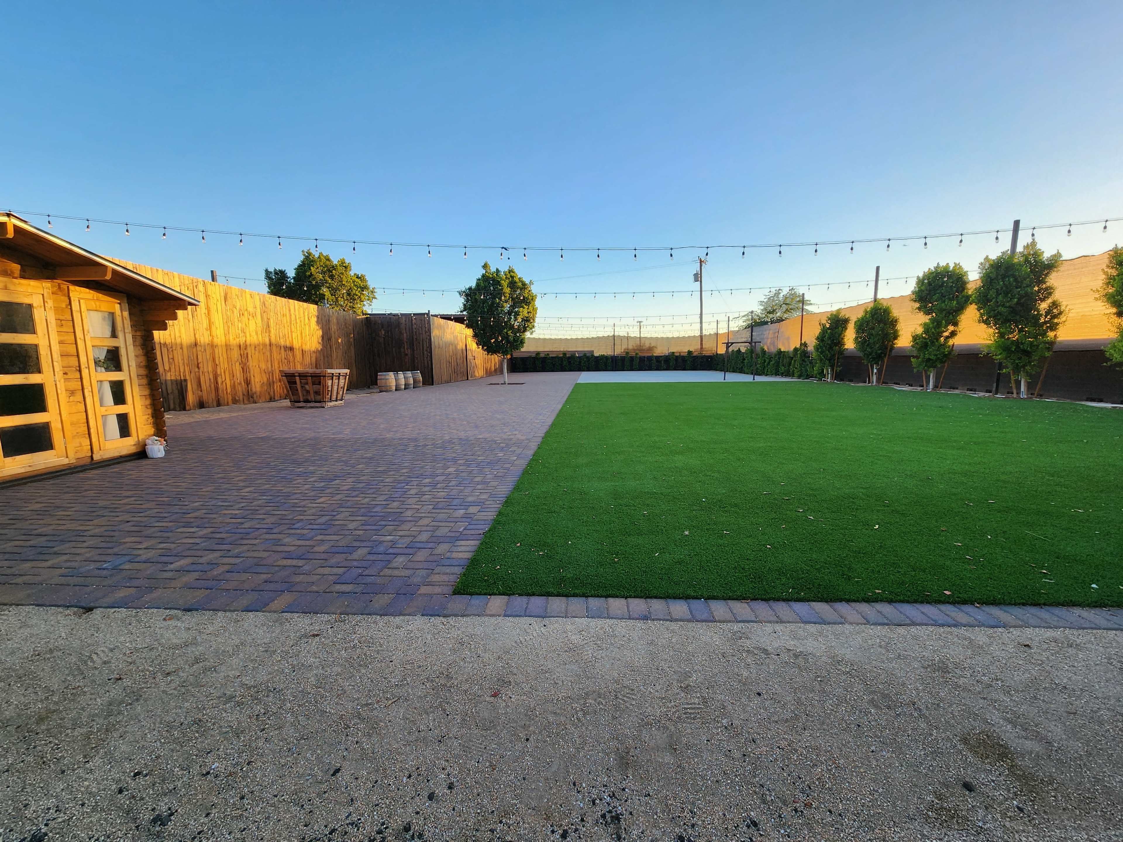 A spacious outdoor area featuring a section of artificial grass, a paved pathway, a wooden shed, and string lights above, with trees lining the perimeter.