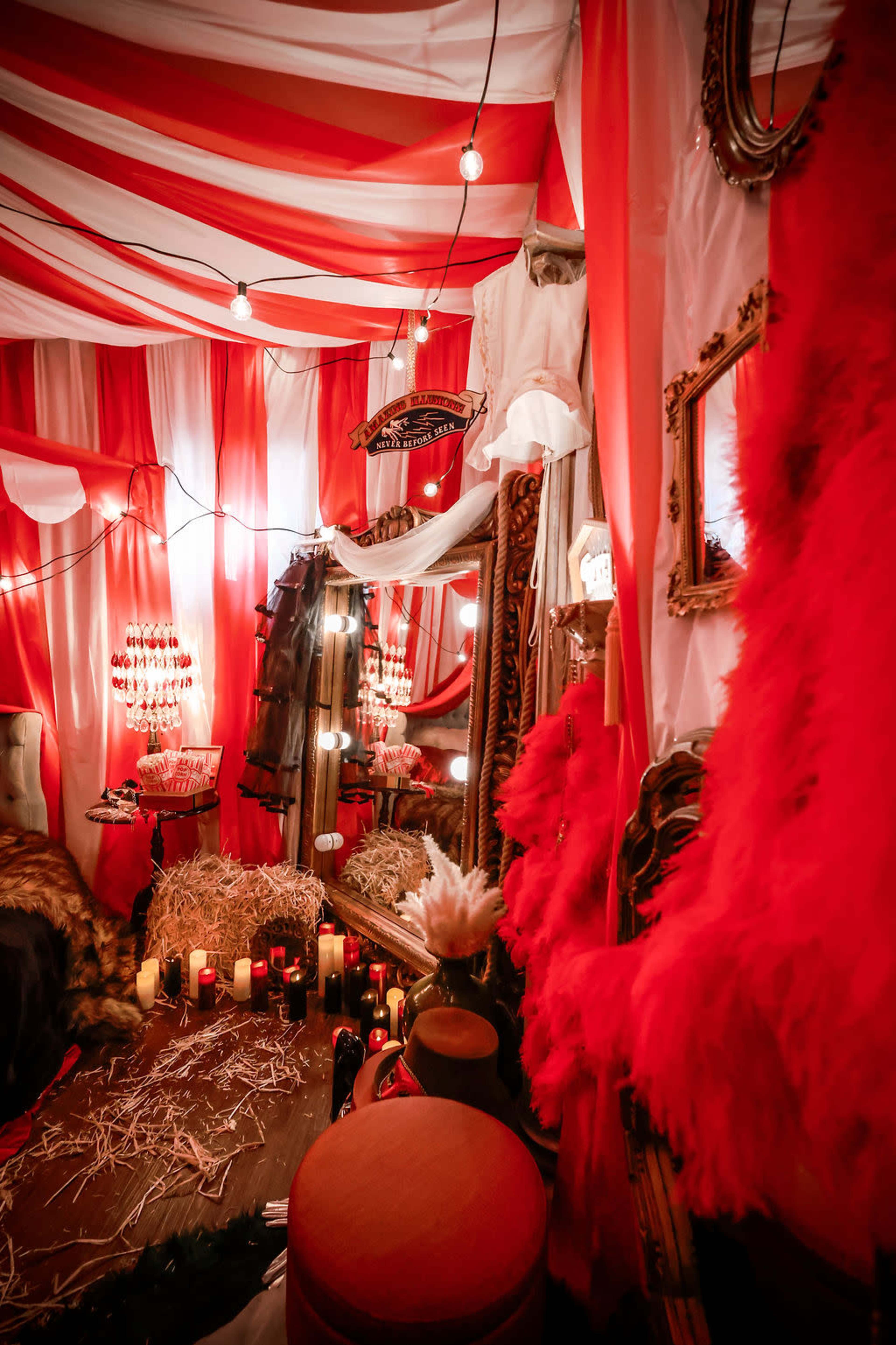 The image shows a vibrant room decorated with red and white fabric draping the ceiling, surrounded by mirrors, decorative elements, and candles arranged on the floor.