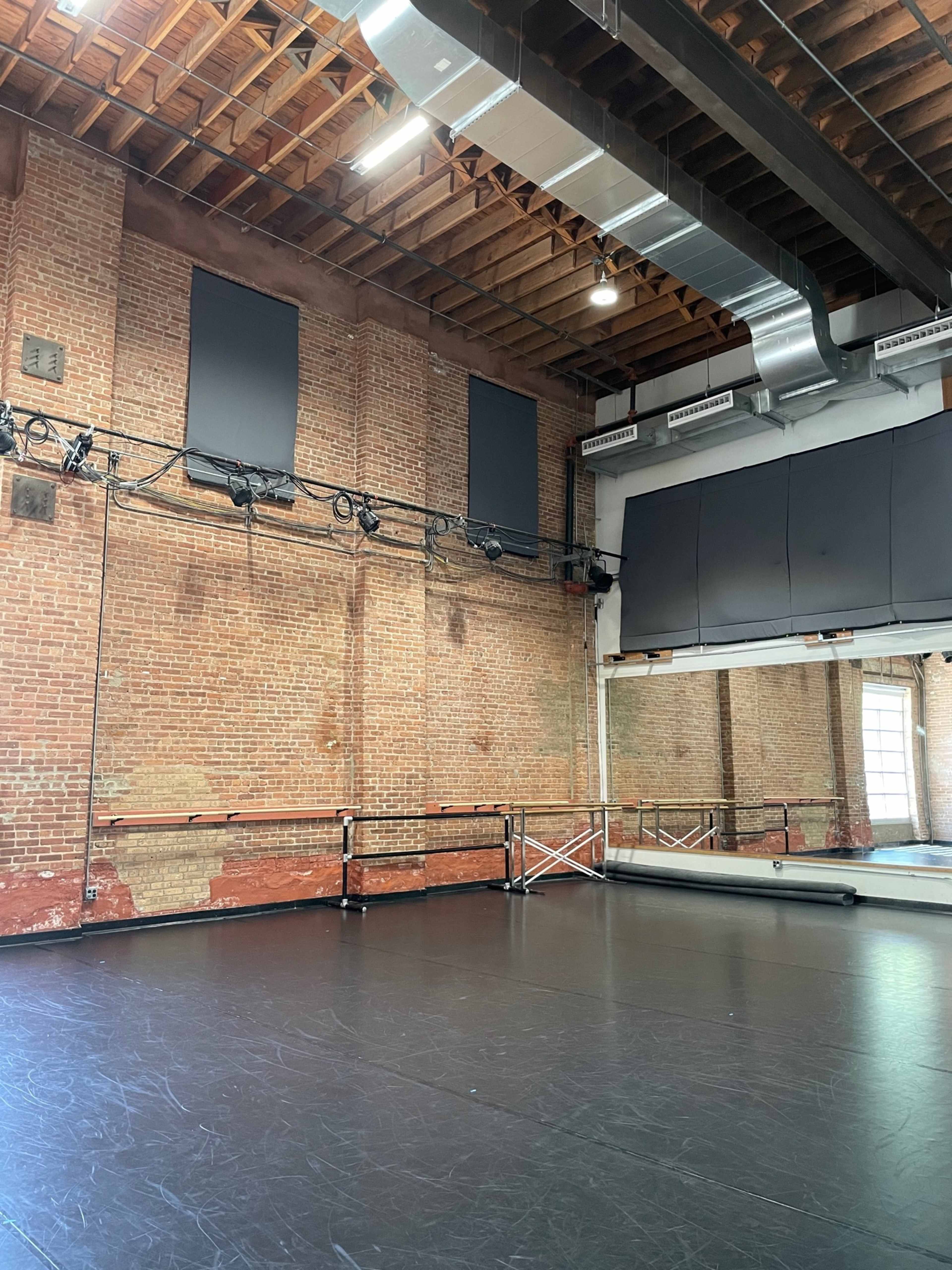 The image shows a spacious dance studio with exposed brick walls, a mirrored wall, and a dark wooden floor.