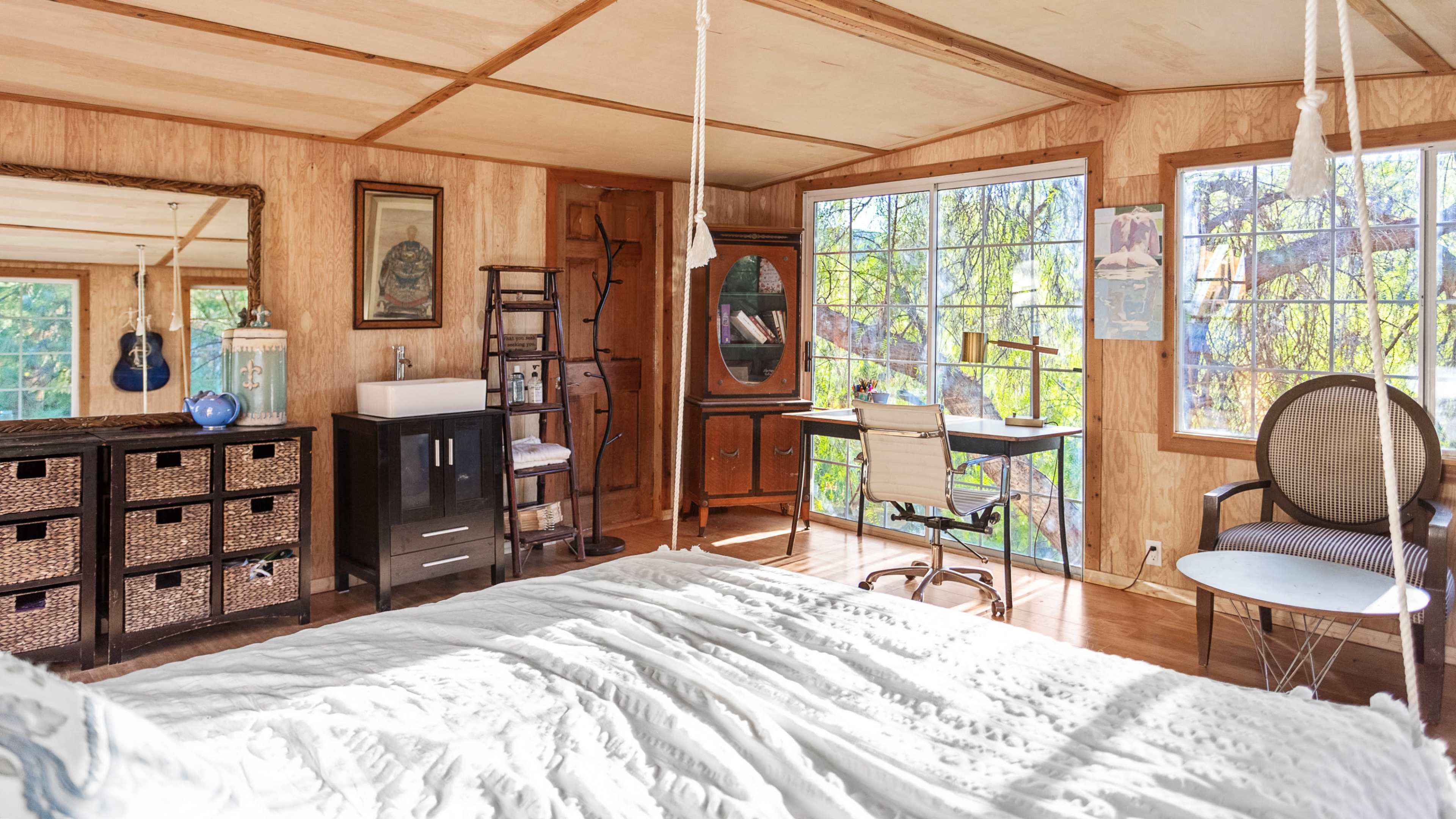 A bright, wooden-floored bedroom featuring a large window, a bed with a white coverlet, a desk with a chair, and storage units.