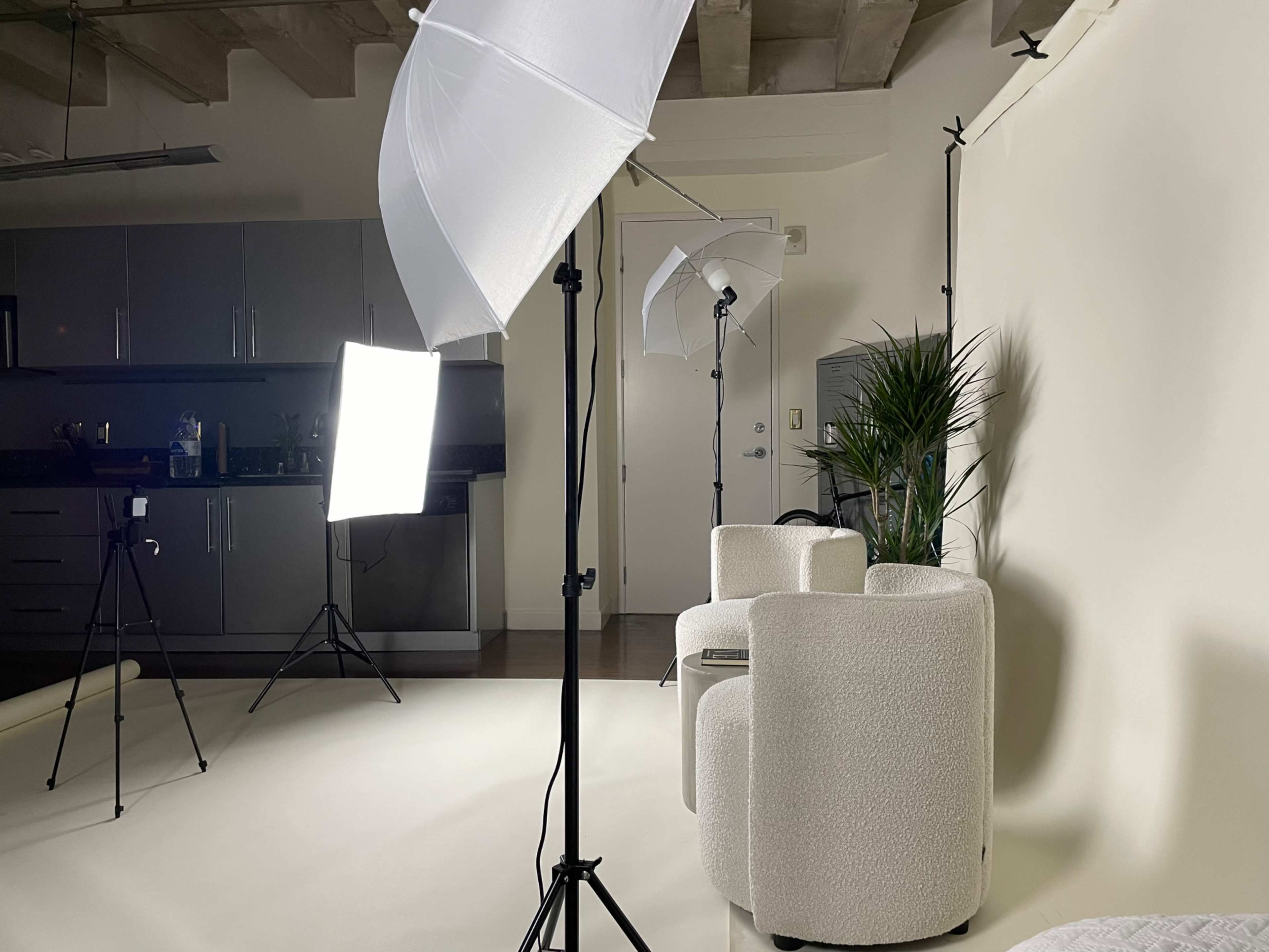 A photography studio setup with two light sources, including umbrella lights, and a minimalist seating arrangement against a plain backdrop.