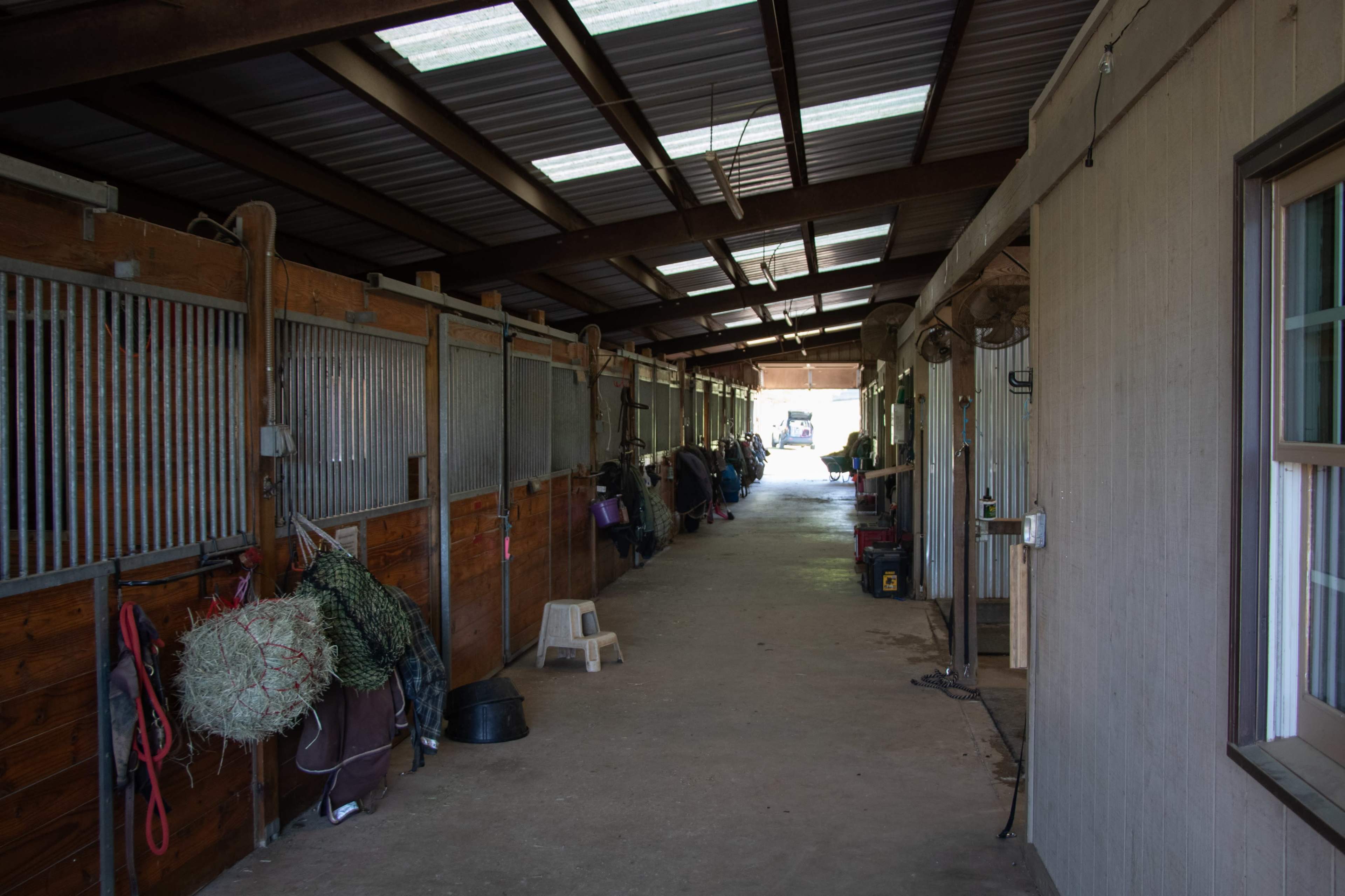 The image shows a long, narrow indoor barn aisle lined with horse stalls on either side.