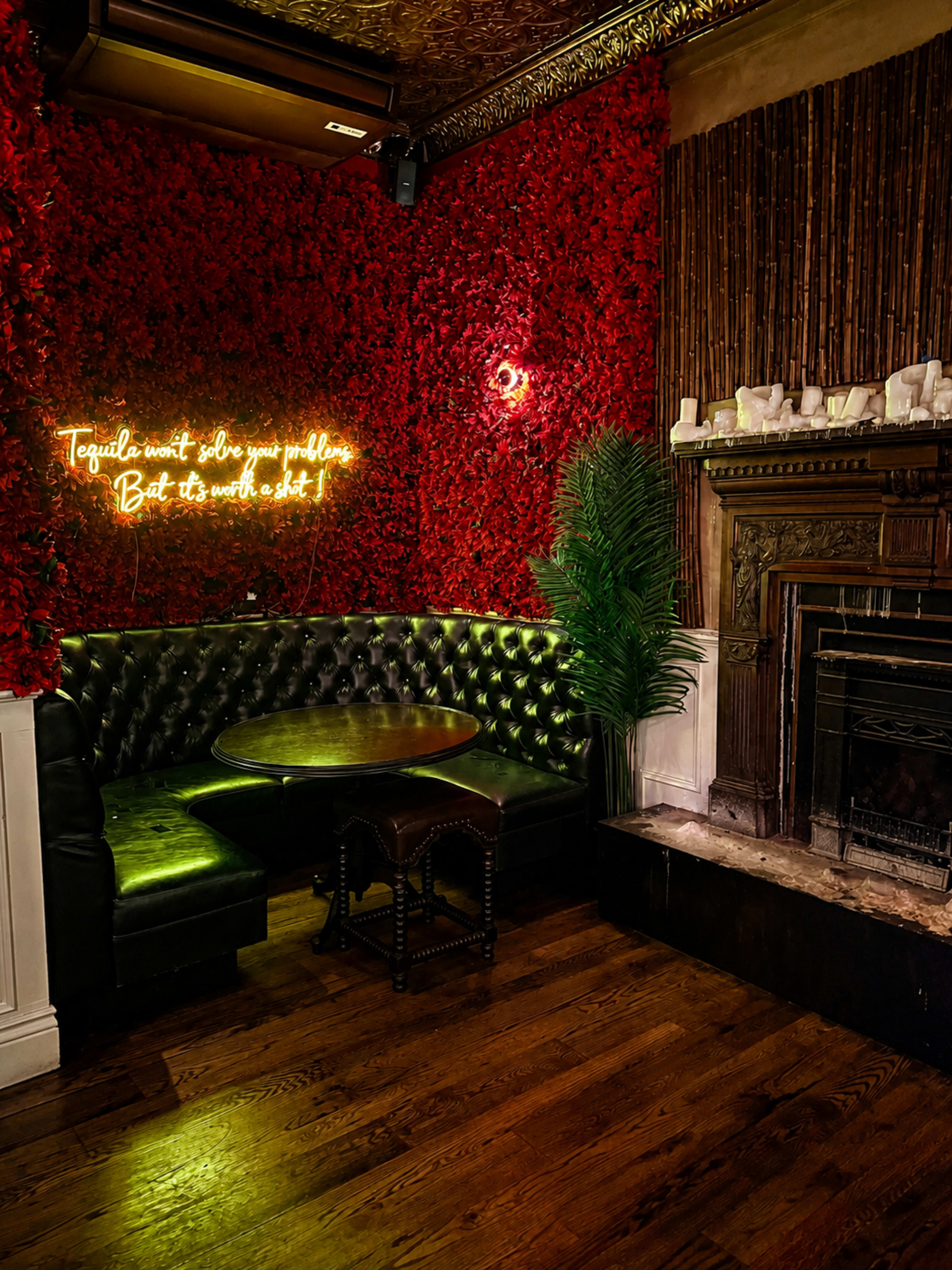 The image shows a cozy seating area in a dimly lit interior, featuring a black leather tufted bench against a wall adorned with red floral decorations, and a vintage fireplace nearby.
