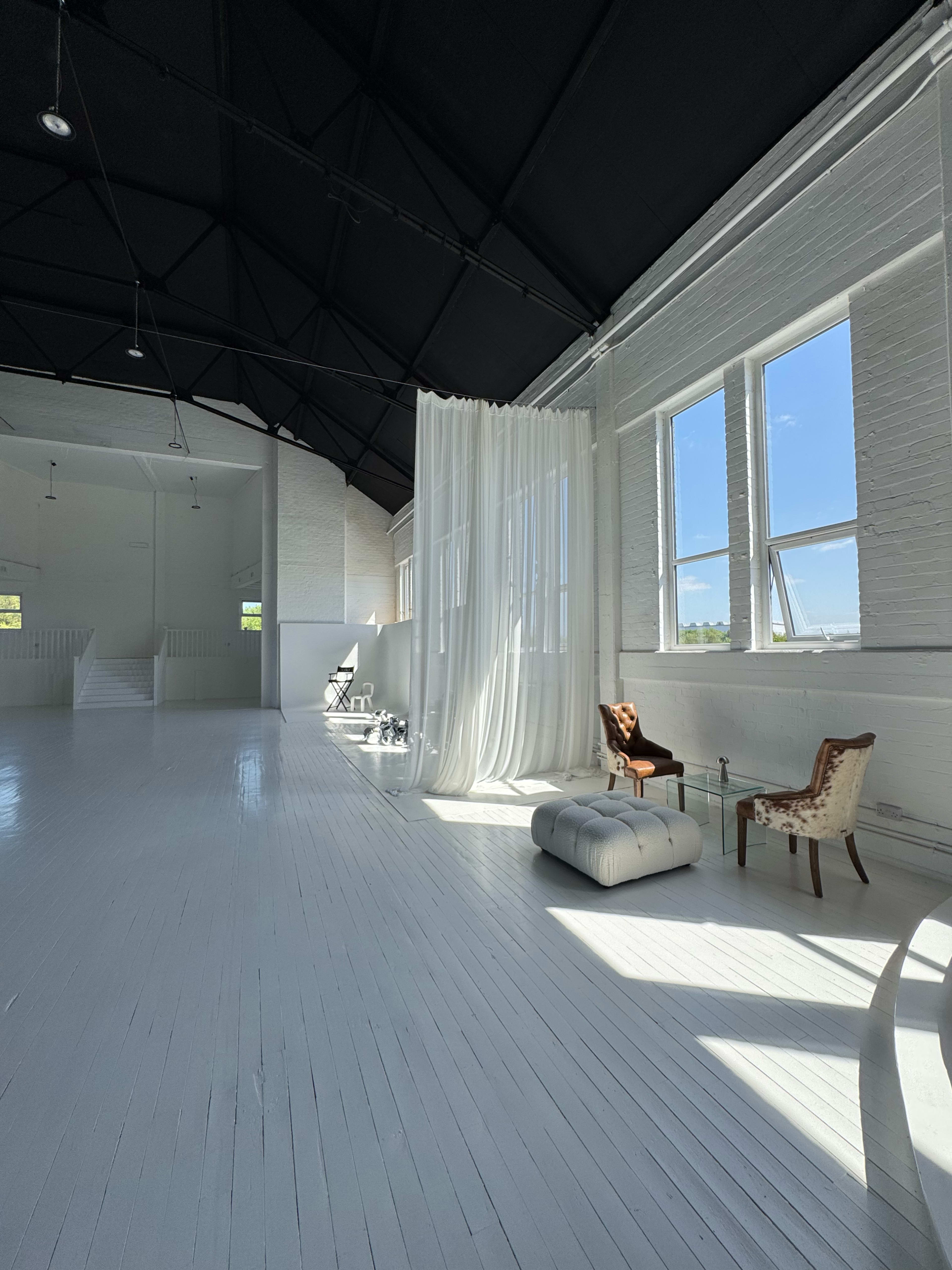 The image shows a spacious, brightly lit room with white walls and floor, featuring large windows, sheer curtains, and a few pieces of furniture.