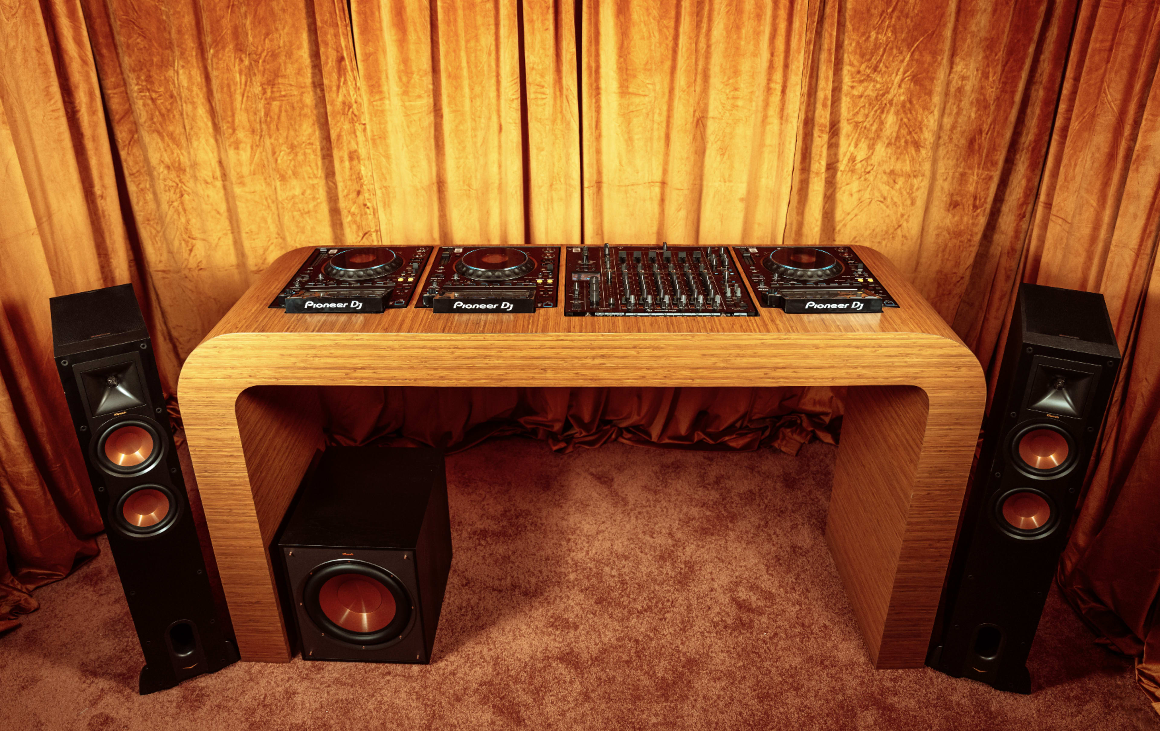 A wooden DJ booth with multiple Pioneer CDJs and a mixer is positioned in front of two large speakers and a subwoofer, against an orange curtain backdrop.