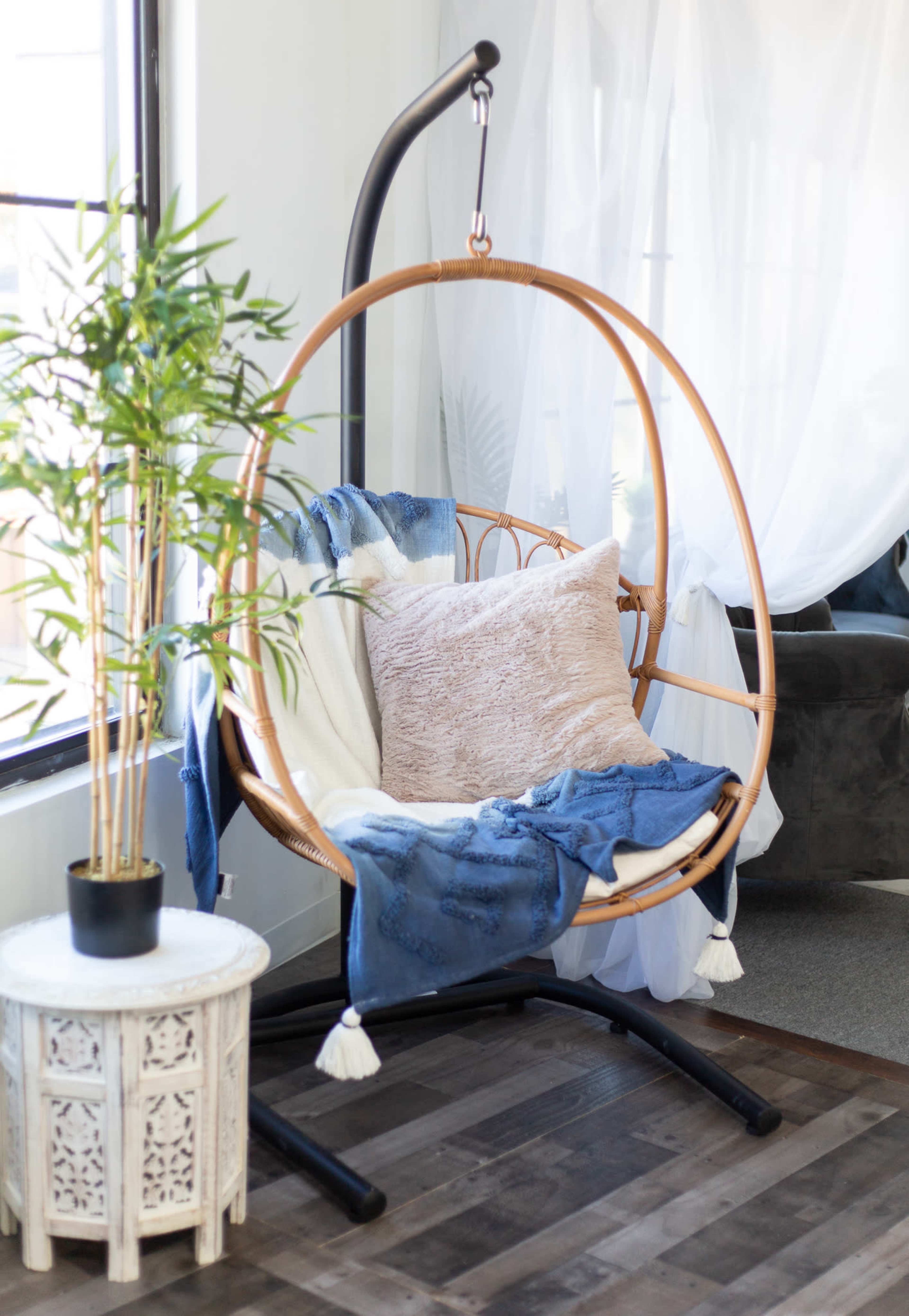 A round woven hanging chair is situated next to a potted plant, draped with a blue blanket and a pillow, beside a decorative side table.