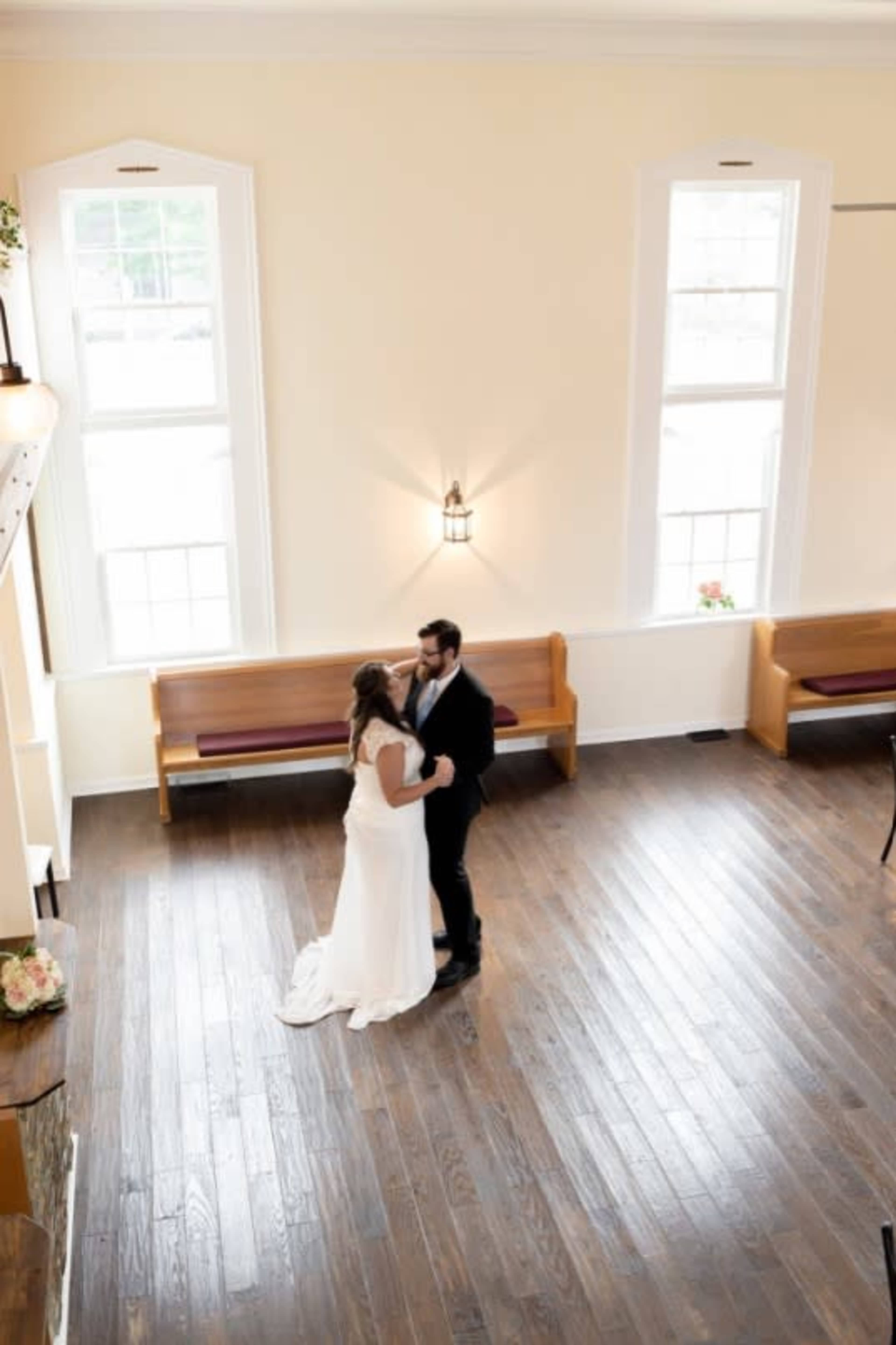 A couple embraces in the center of a bright, spacious room with wooden floors and simple pews.