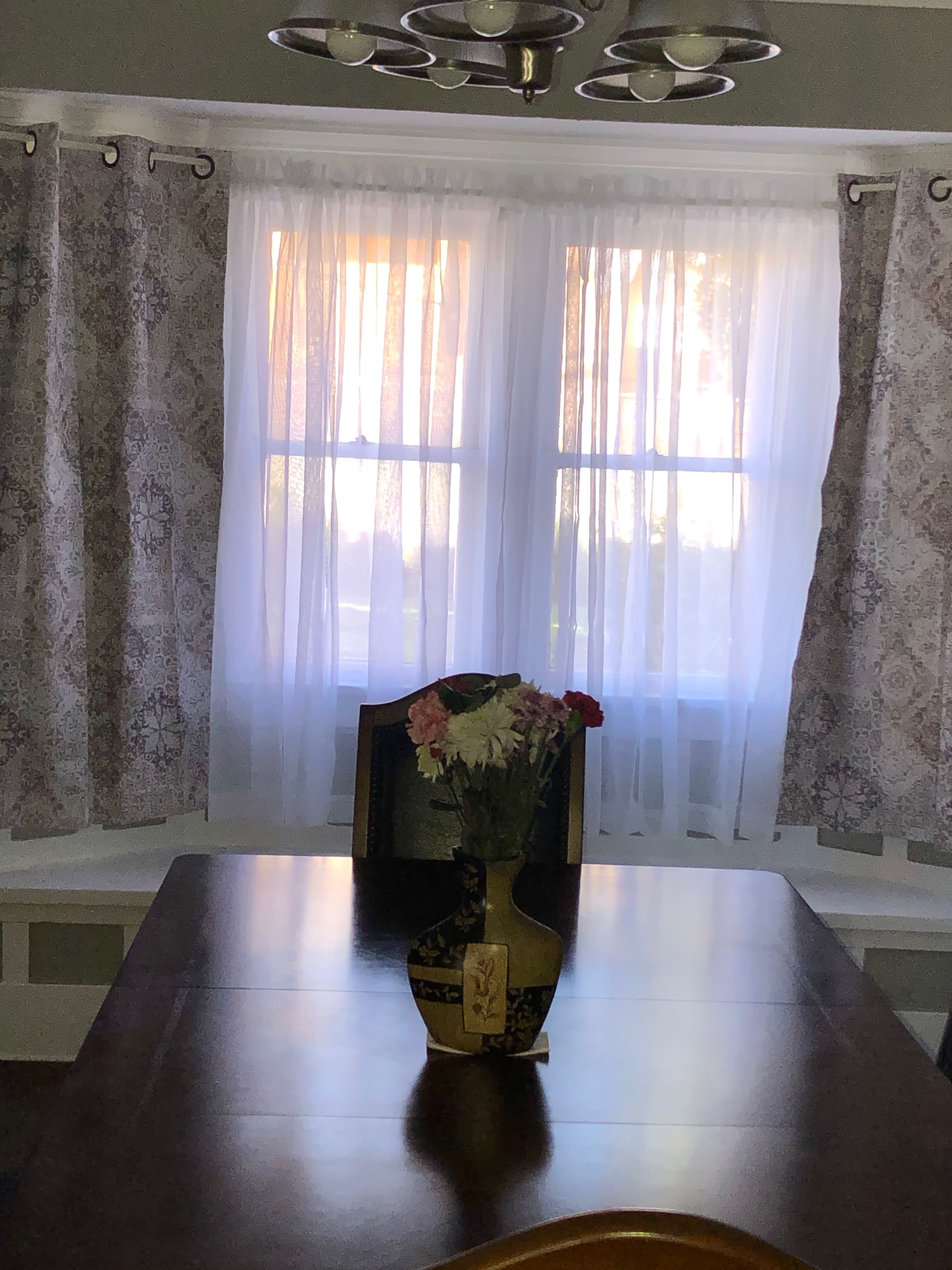 A dining area features a wooden table with a vase of flowers at its center, surrounded by two windows dressed with sheer and patterned curtains.