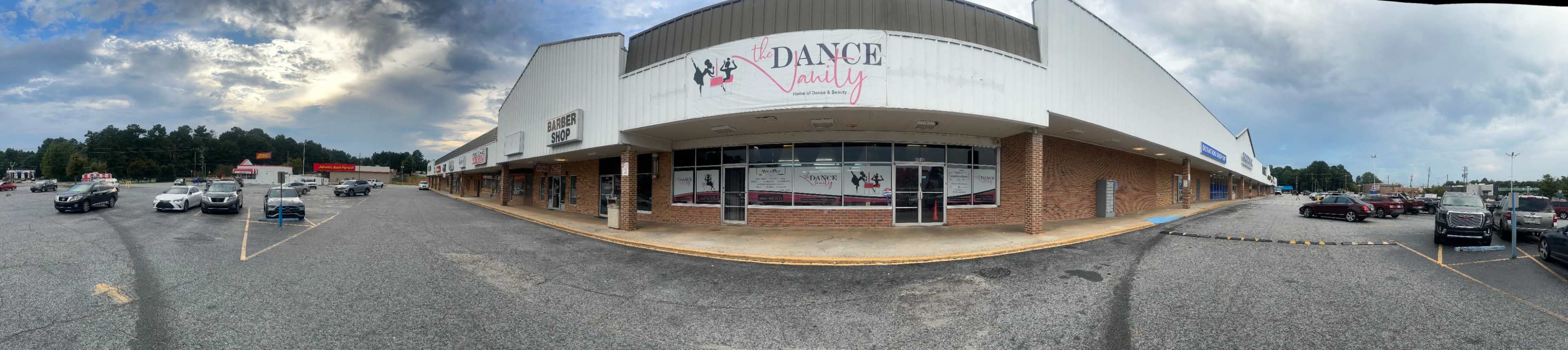 The image shows a panoramic view of a shopping plaza featuring a dance studio with large windows and a sign in front, surrounded by a parking lot filled with cars.