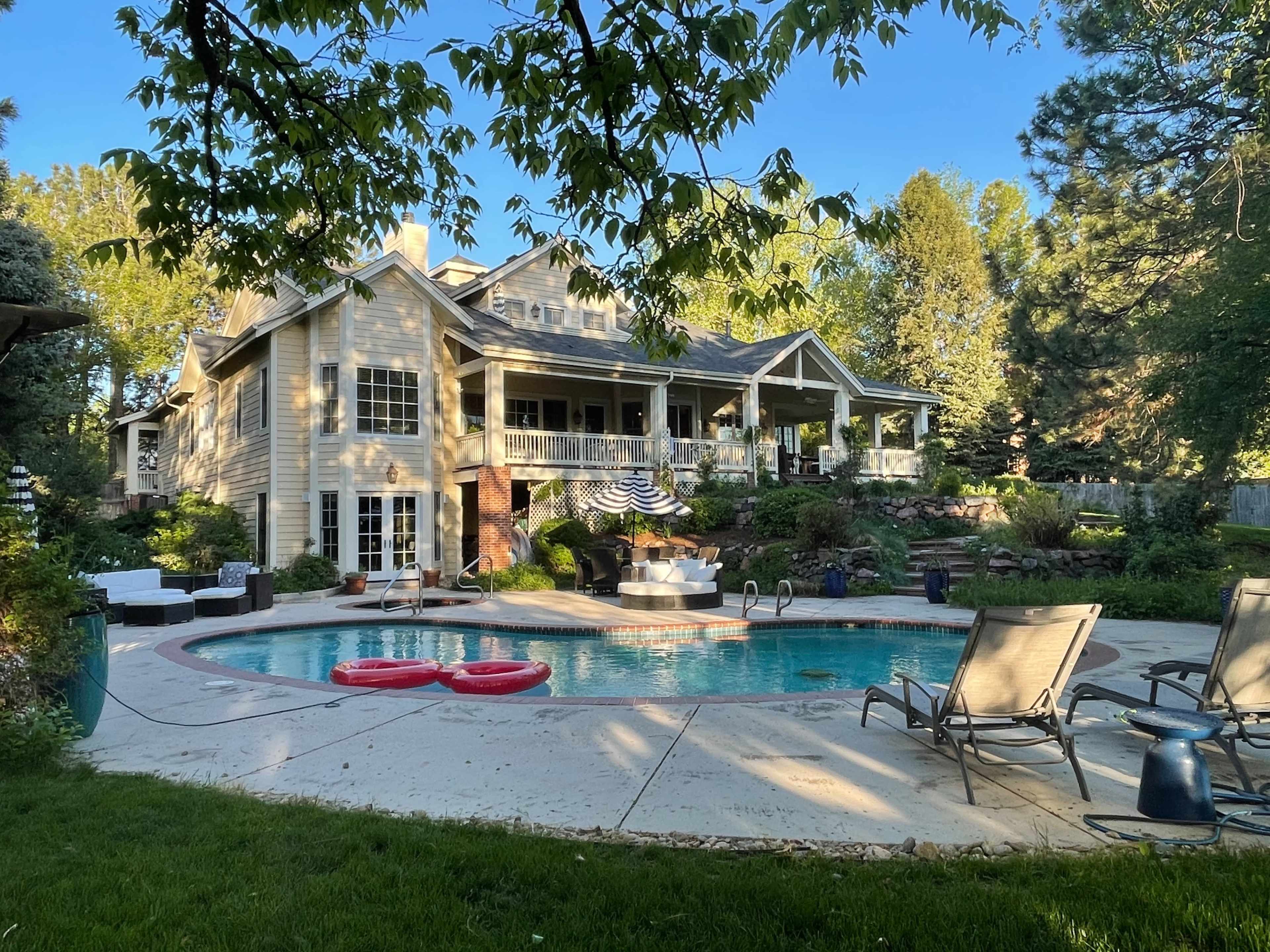 A spacious backyard features a swimming pool surrounded by lounge chairs and floats, with a large house and landscaped greenery in the background.