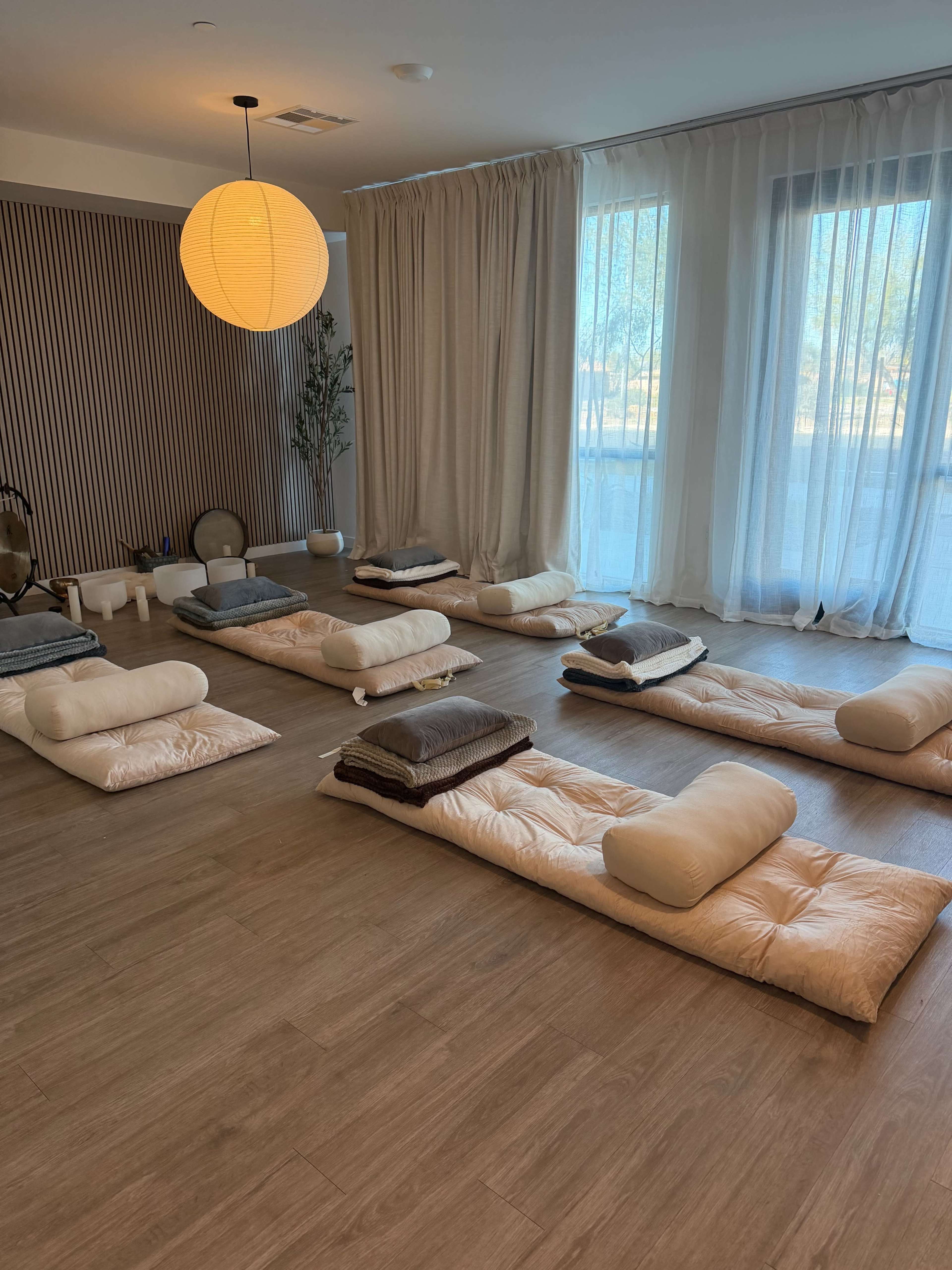 A spacious wellness room features several neatly arranged floor mats and cushions under soft lighting and a large window.