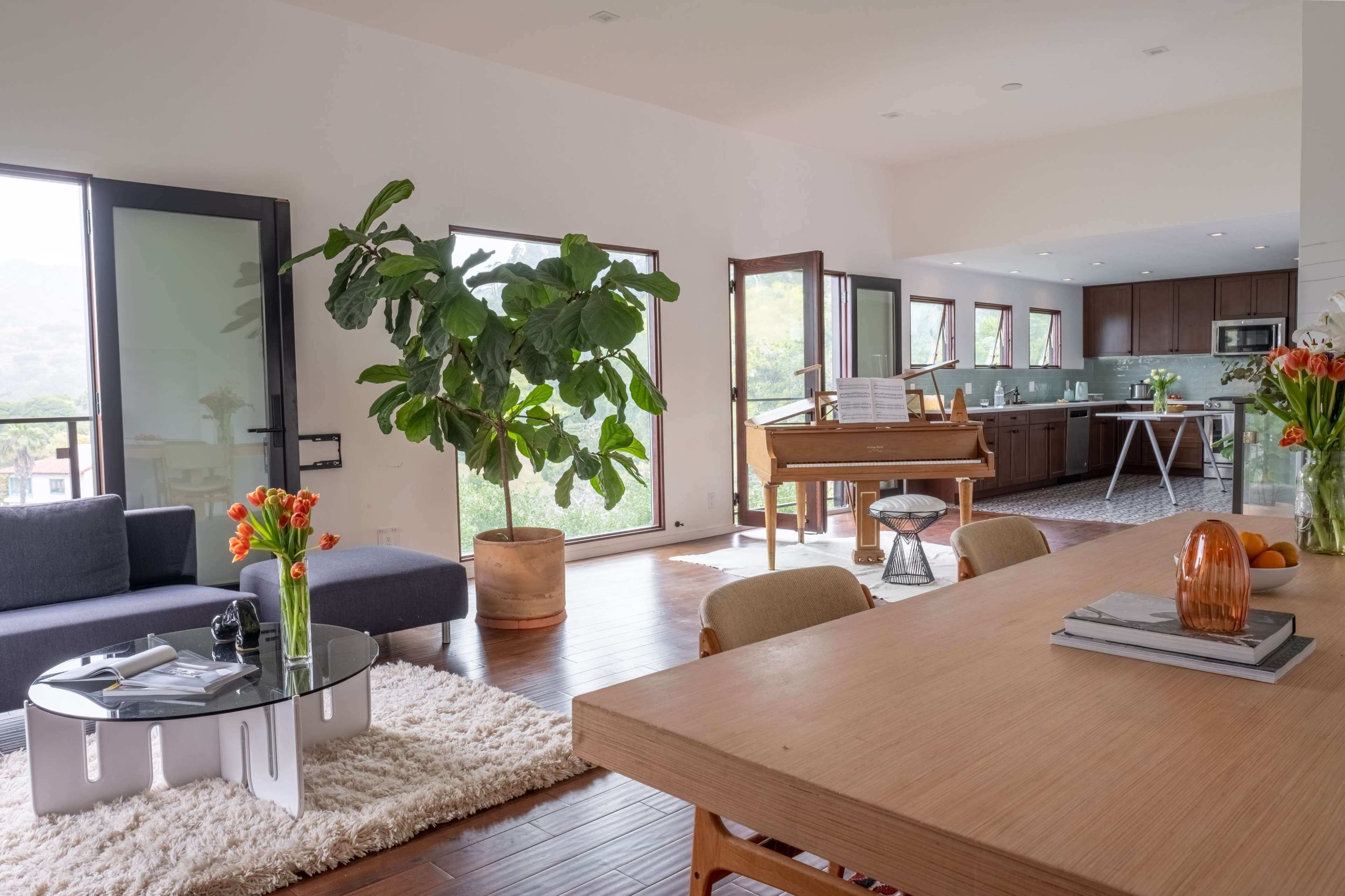 A spacious living area with a modern sofa, a wooden dining table, and a piano, featuring large windows that provide natural light and a view of the outdoors.
