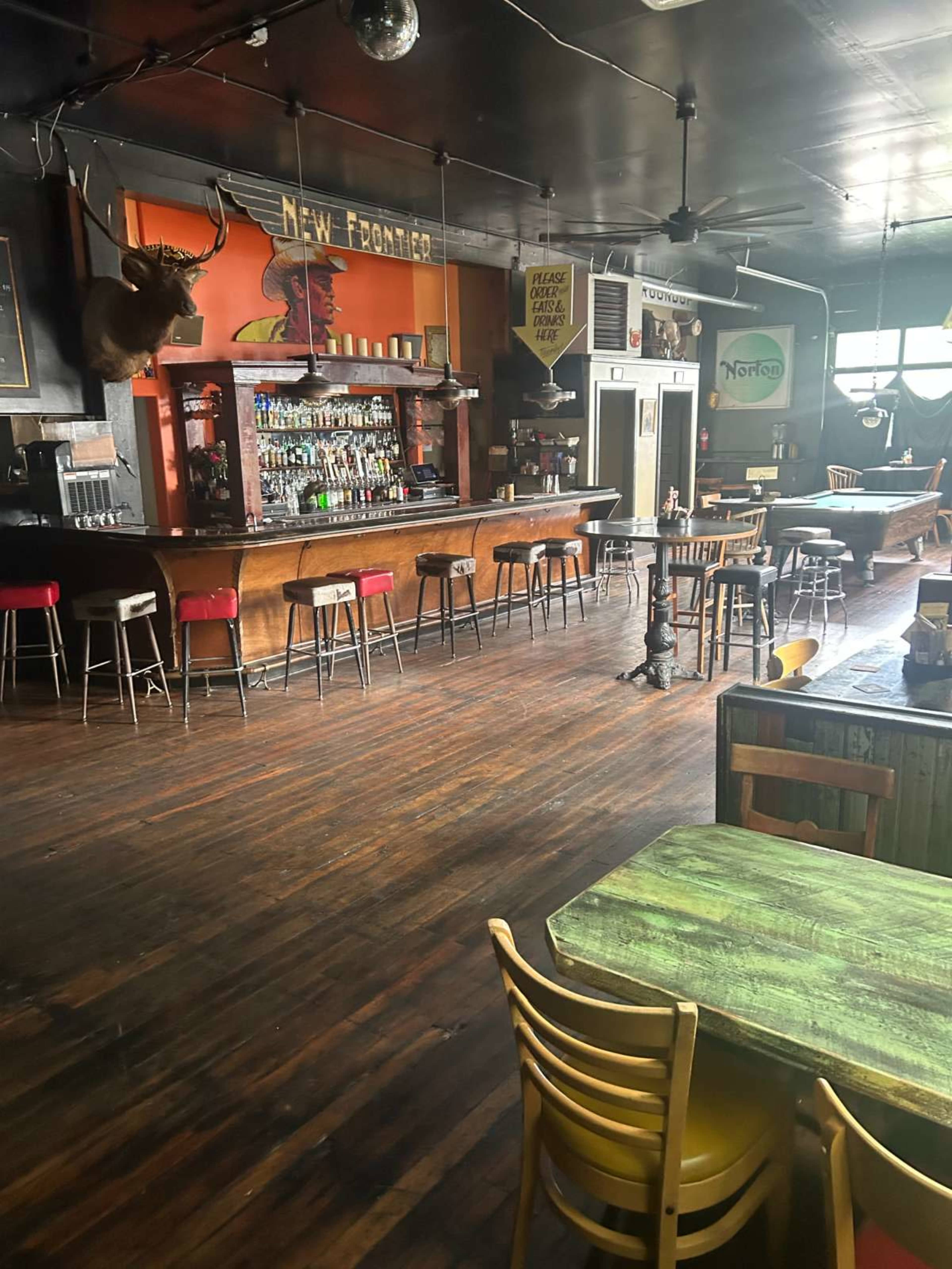 The image shows a spacious bar interior with a long wooden counter, several high chairs, a deer head mount on the wall, and various tables scattered around the room.
