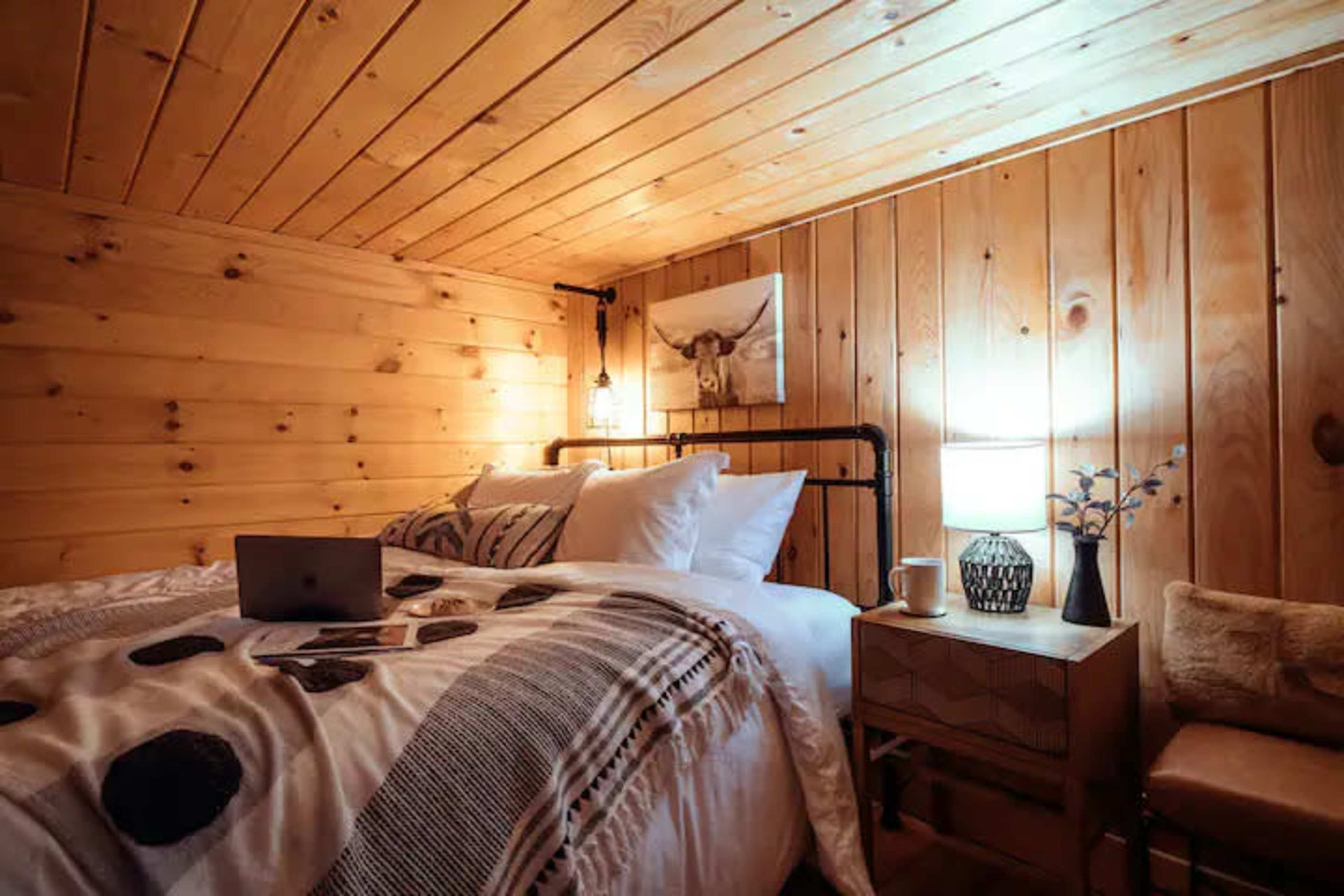 A cozy bedroom features a wooden wall, a neatly made bed with decorative pillows, a bedside table with a lamp and a cup, and a laptop on the bed.