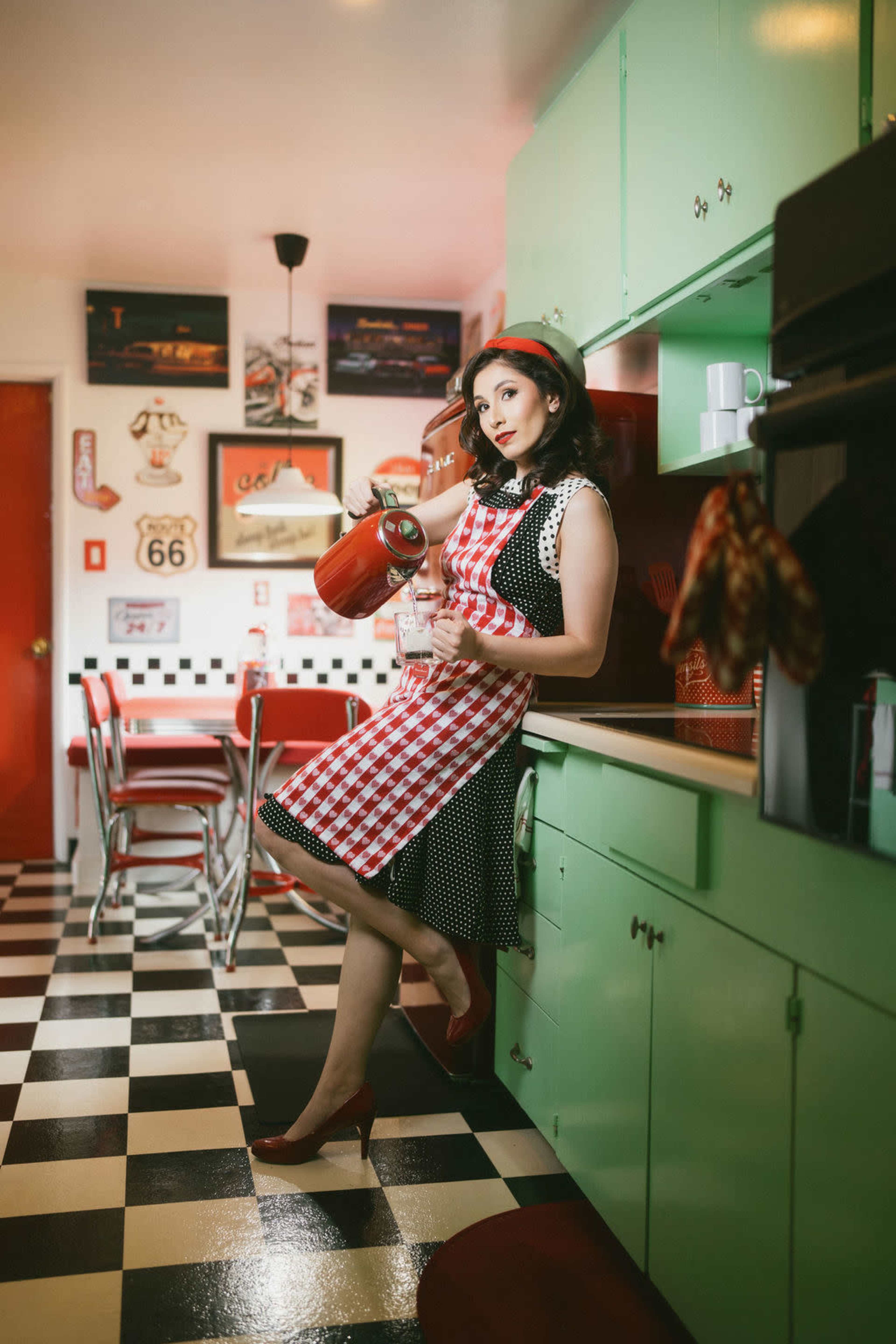A woman in a vintage kitchen wears a red and white checkered dress and holds a red fire extinguisher while leaning casually against a green cabinet.