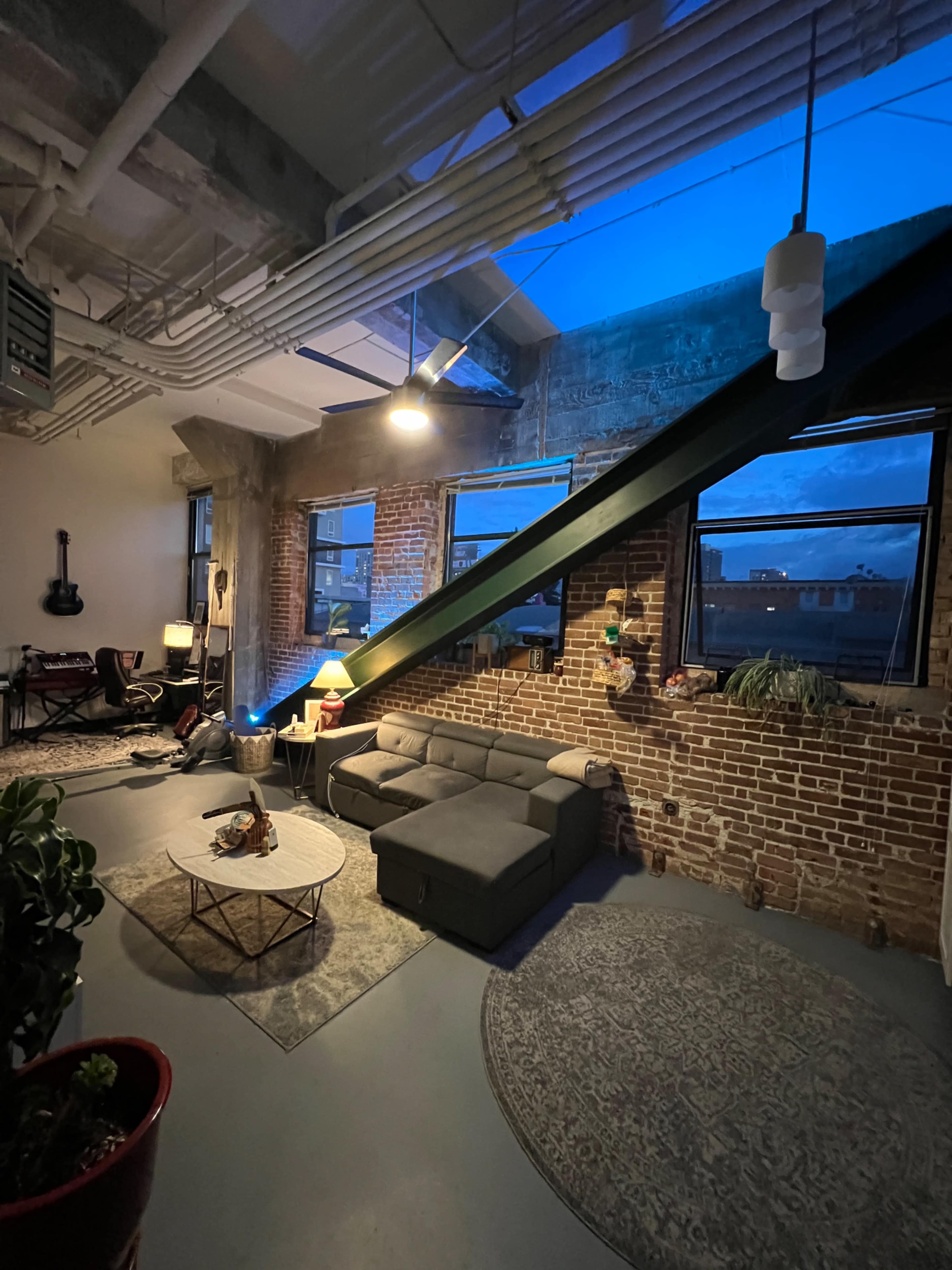 The image shows a modern living room with exposed brick walls, large windows, and a slanted beam, featuring a gray sectional sofa, a coffee table, and decorative plants.