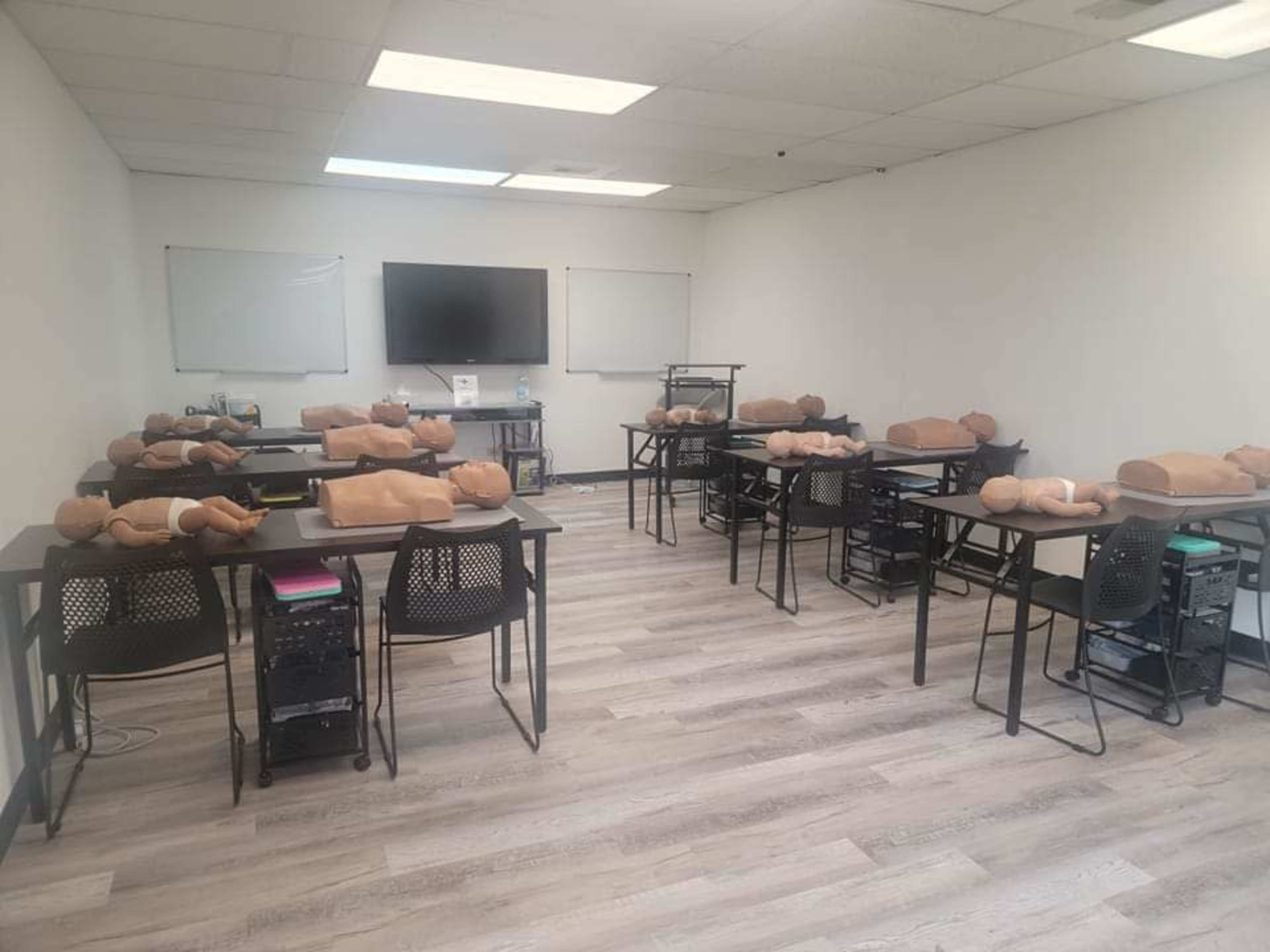 A classroom is set up with several tables, each featuring a lifelike training mannequin, and monitors are positioned at the front of the room.