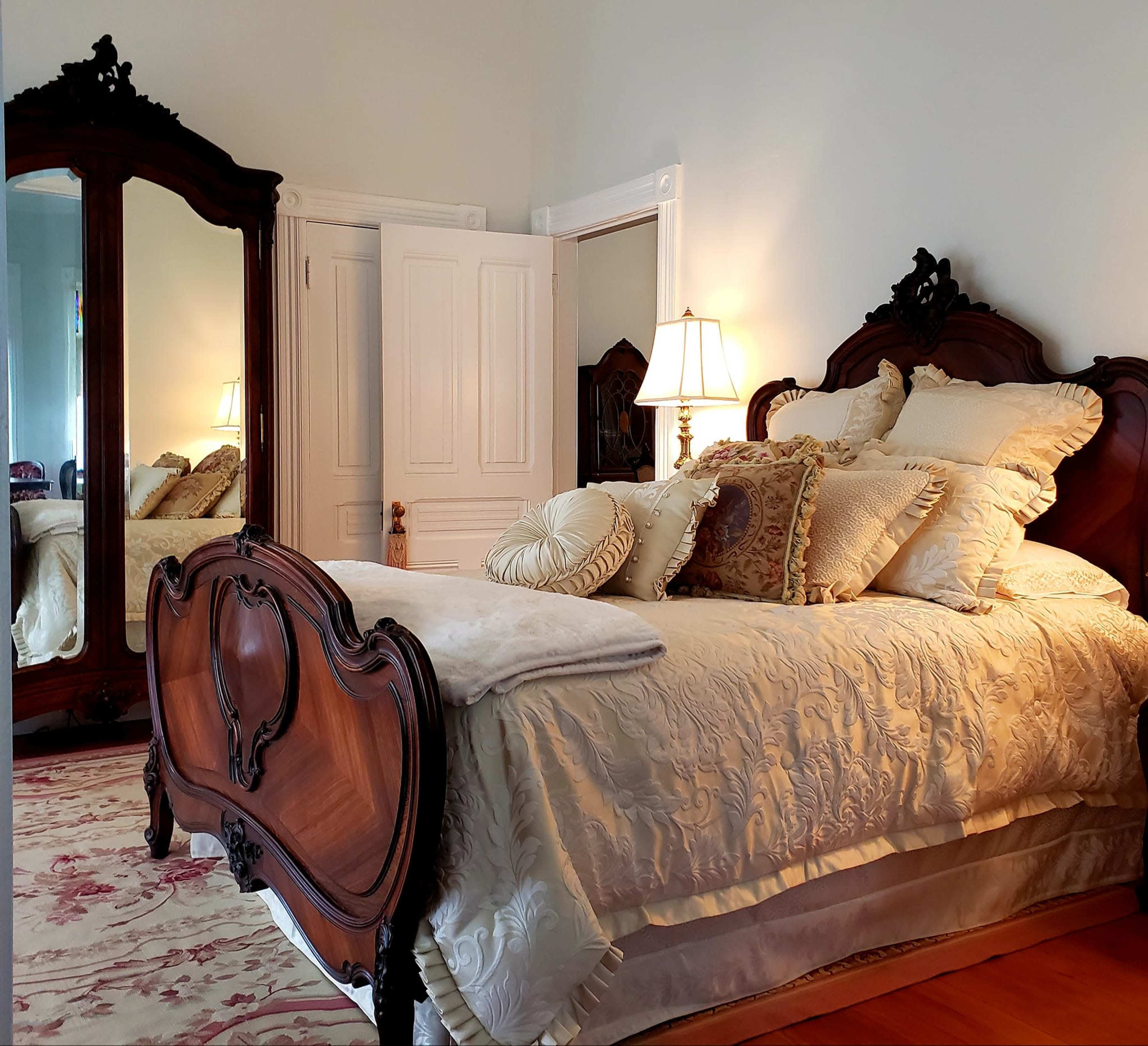 The image shows a well-decorated bedroom featuring a large bed with decorative pillows, an ornate wooden headboard, a matching mirror, and a bedside lamp.