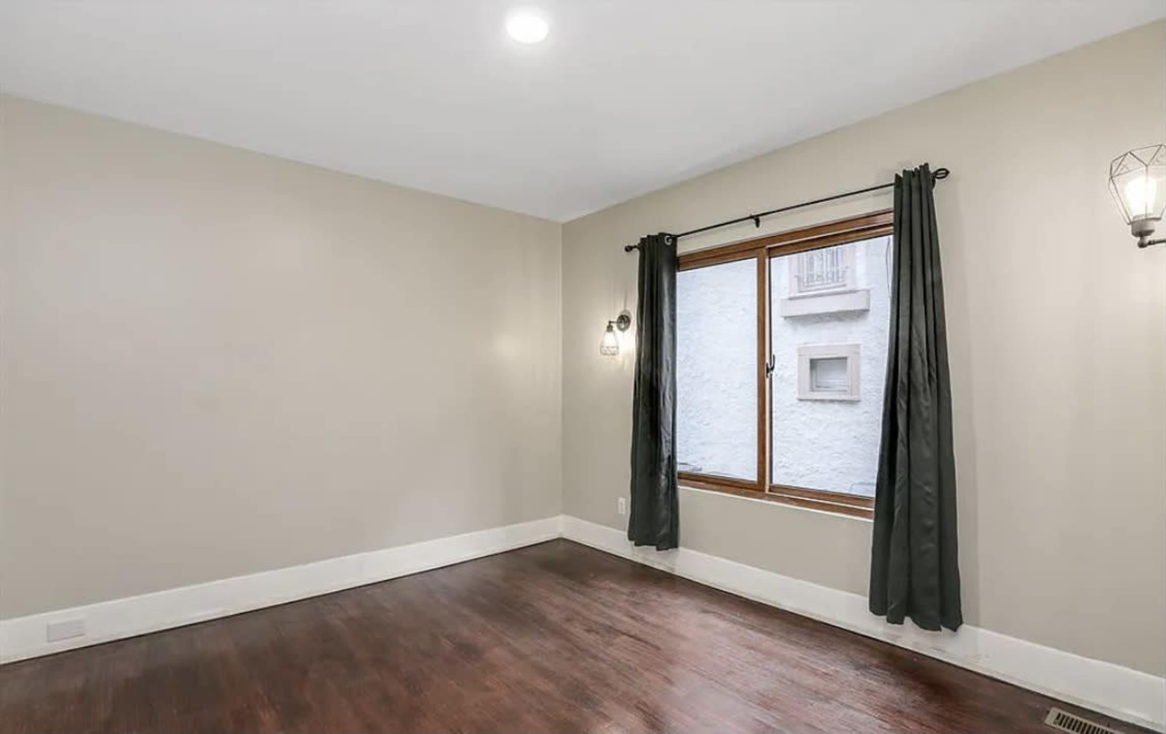 A sparsely furnished room with hardwood flooring and a window framed by dark curtains.