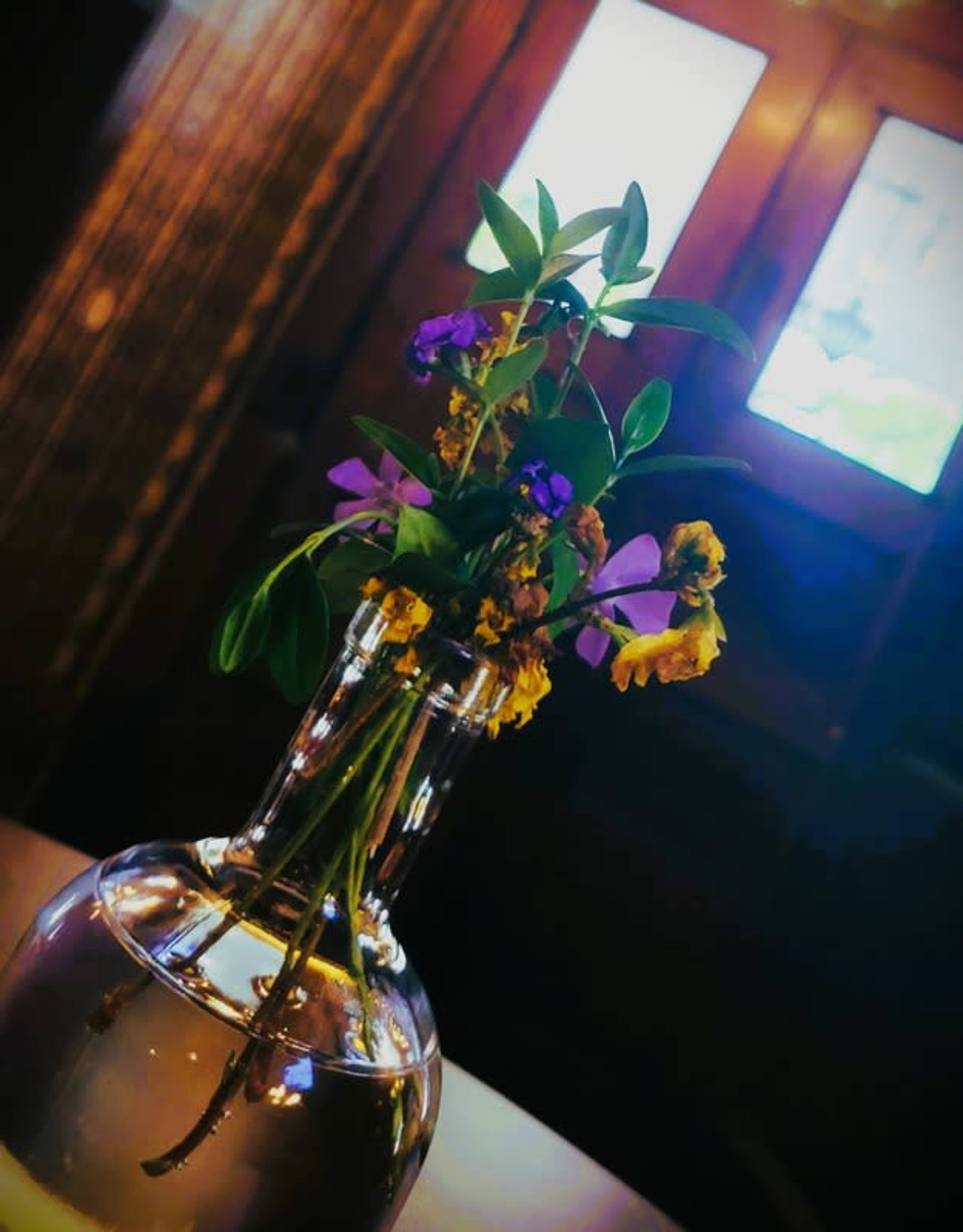 A glass vase holds a small arrangement of colorful flowers with greenery, placed on a table in front of a window.