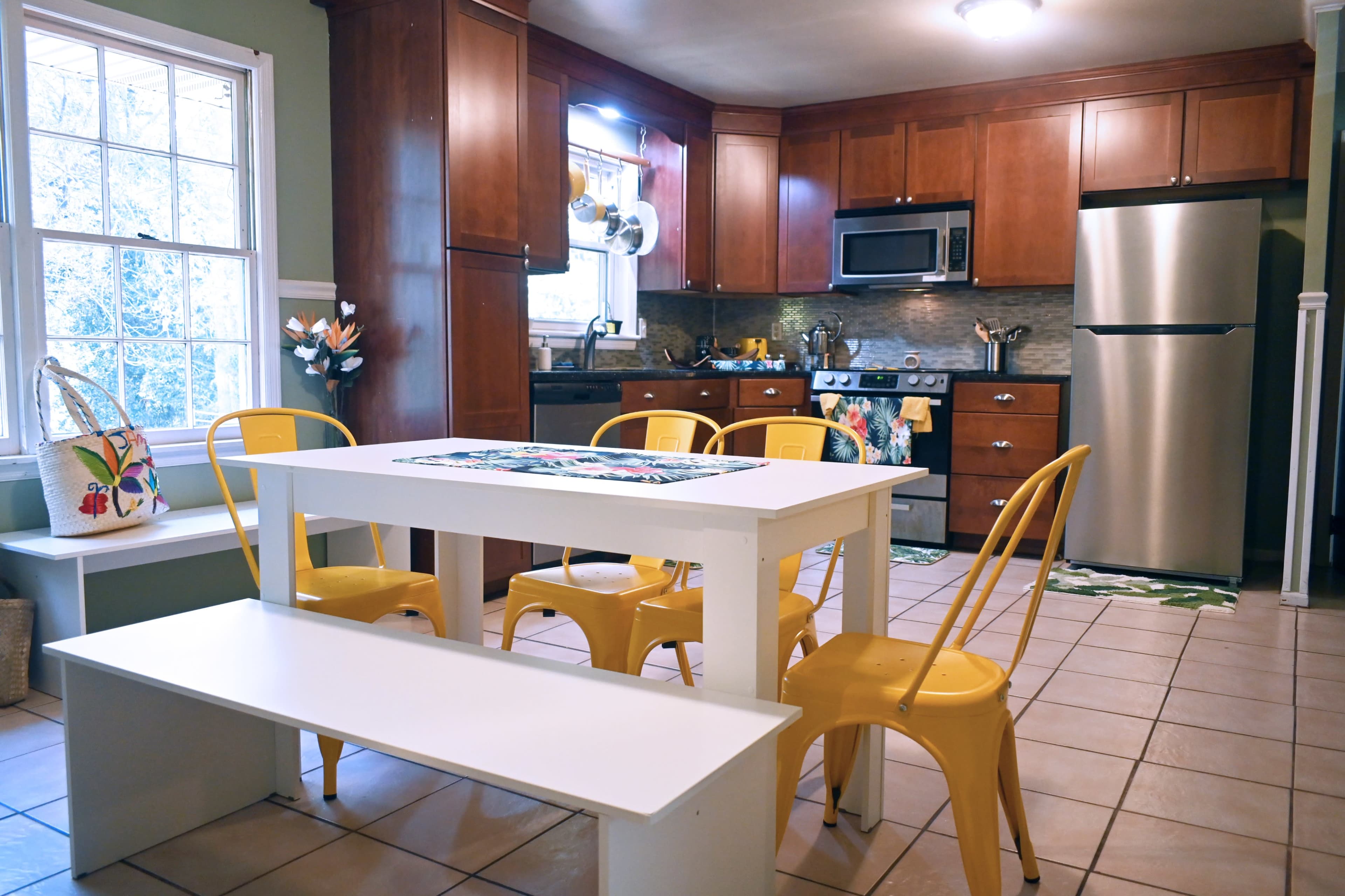 A modern kitchen with wooden cabinetry, stainless steel appliances, and a white dining table surrounded by yellow chairs.